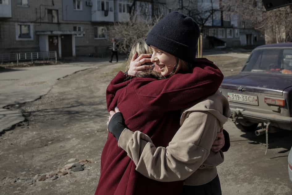 Две женщины, обе в тёплой верхней одежде (одна в бордовом пальто, другая в бежевой толстовке и чёрной шапке), крепко обнимаются на улице. На заднем плане видны старые жилые дома и припаркованный тёмный автомобиль. Атмосфера тёплая и эмоциональная.