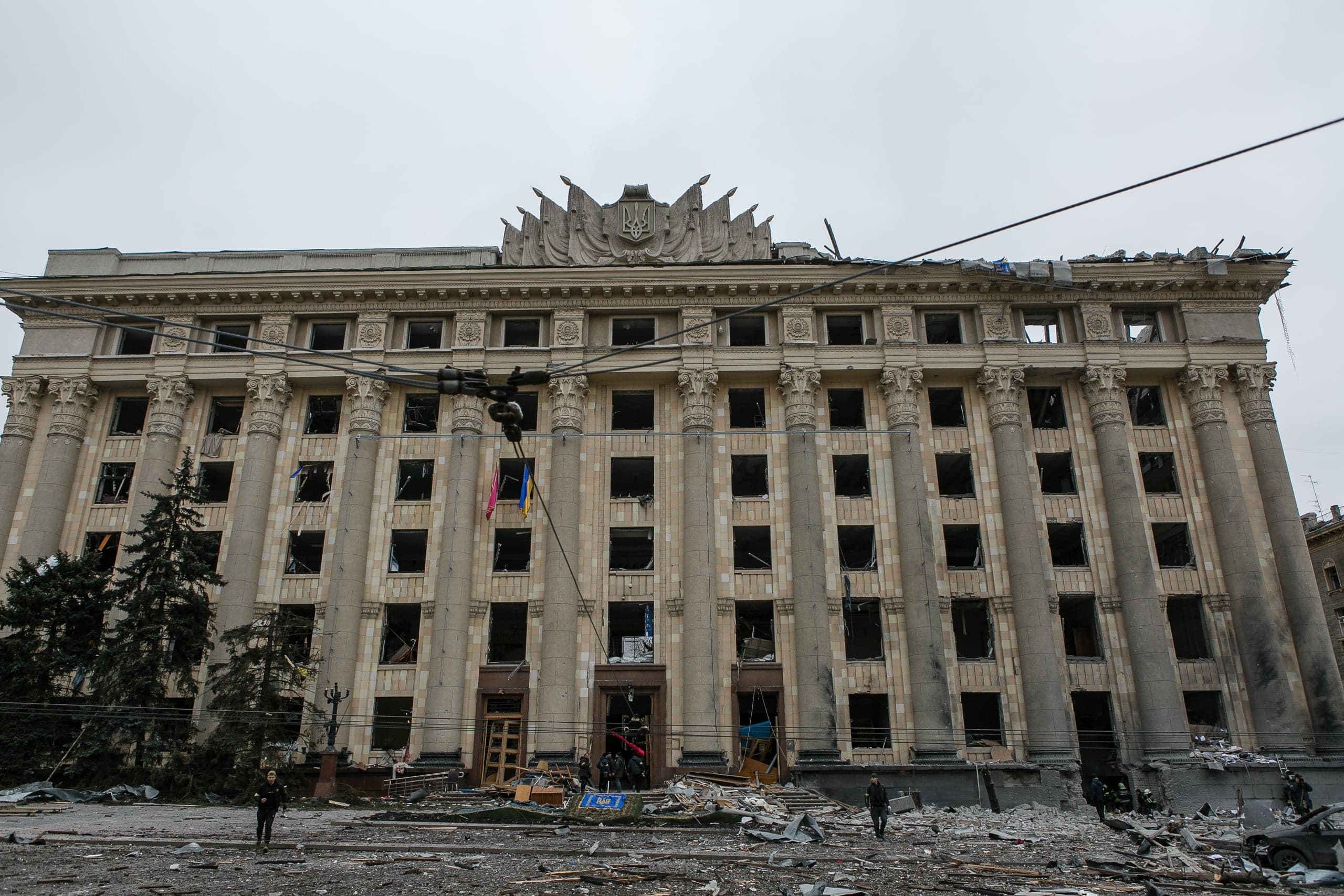 Разрушенное здание Харьковской областной администрации с выбитыми окнами, поврежденной крышей и обломками на площади перед ним. Несколько человек на фоне.
