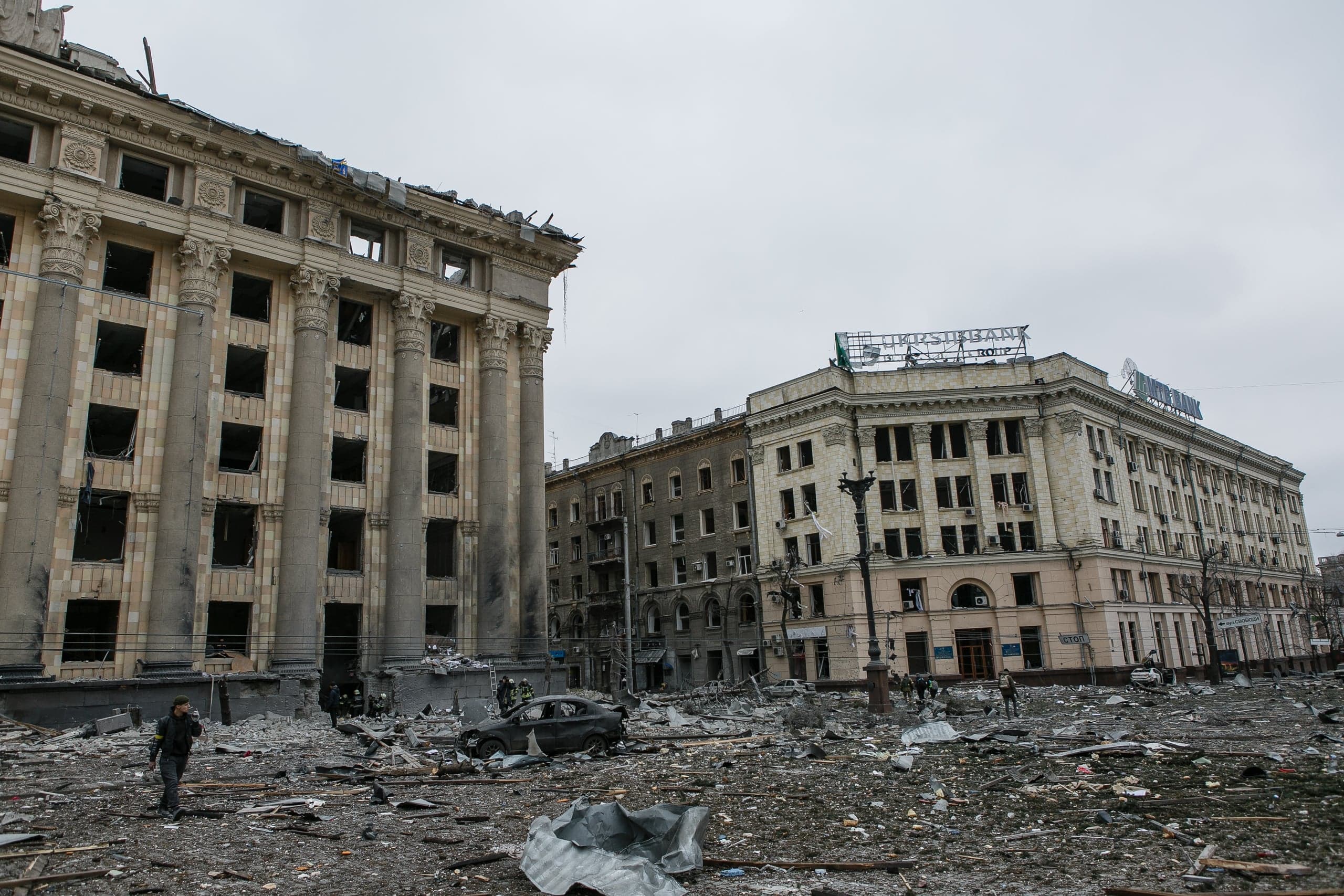 Разрушенные бомбардировкой здания в стиле неоклассицизма, с выбитыми окнами и обломками на площади; видны остатки машин и люди. На здании справа надпись "УКРСИББАНК".