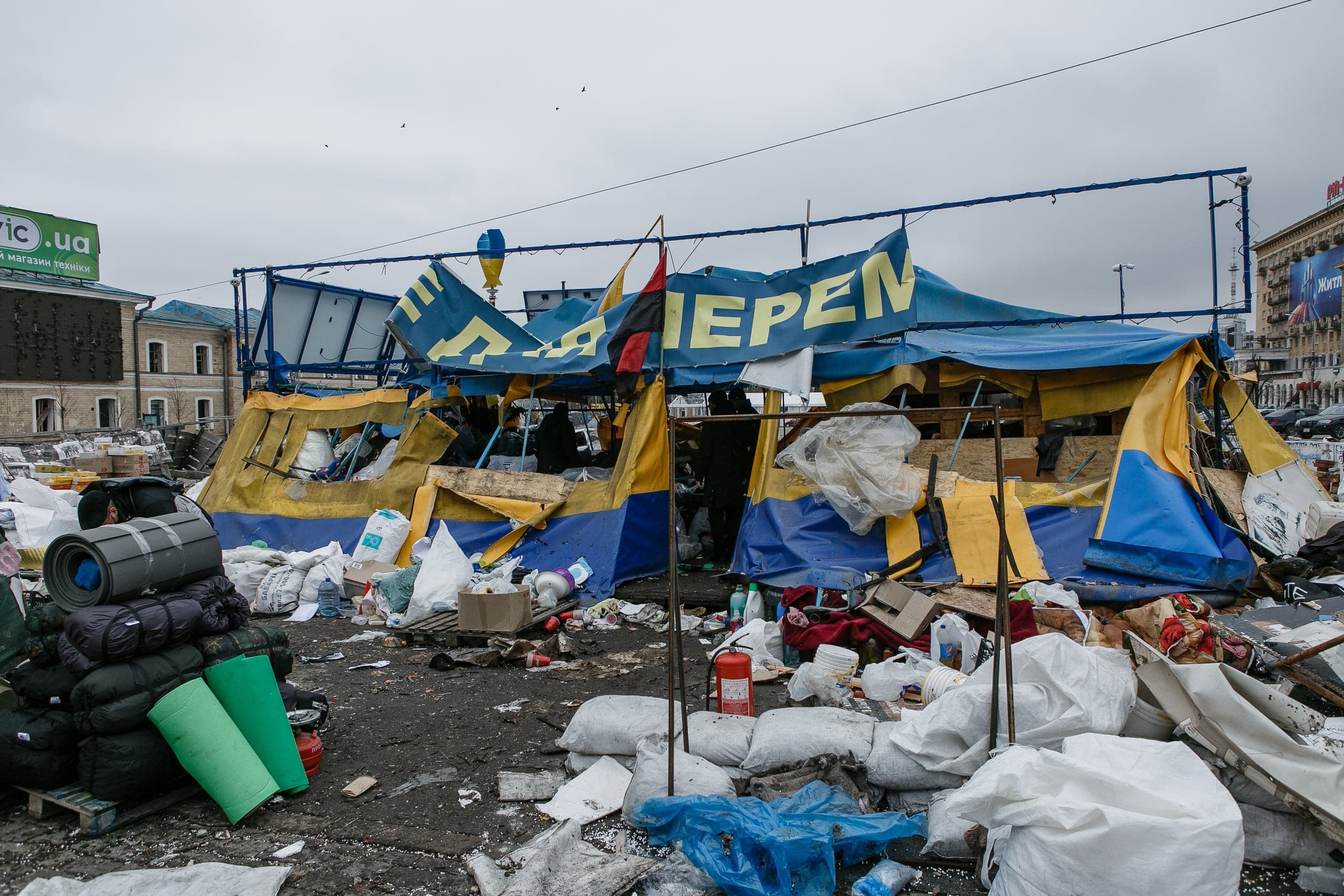 Временный лагерь из синих и желтых тарпов, окруженный мусором, мешками и туристическими принадлежностями. На баннере виден желтый текст, фрагмент "ПЕРЕПЕН".
