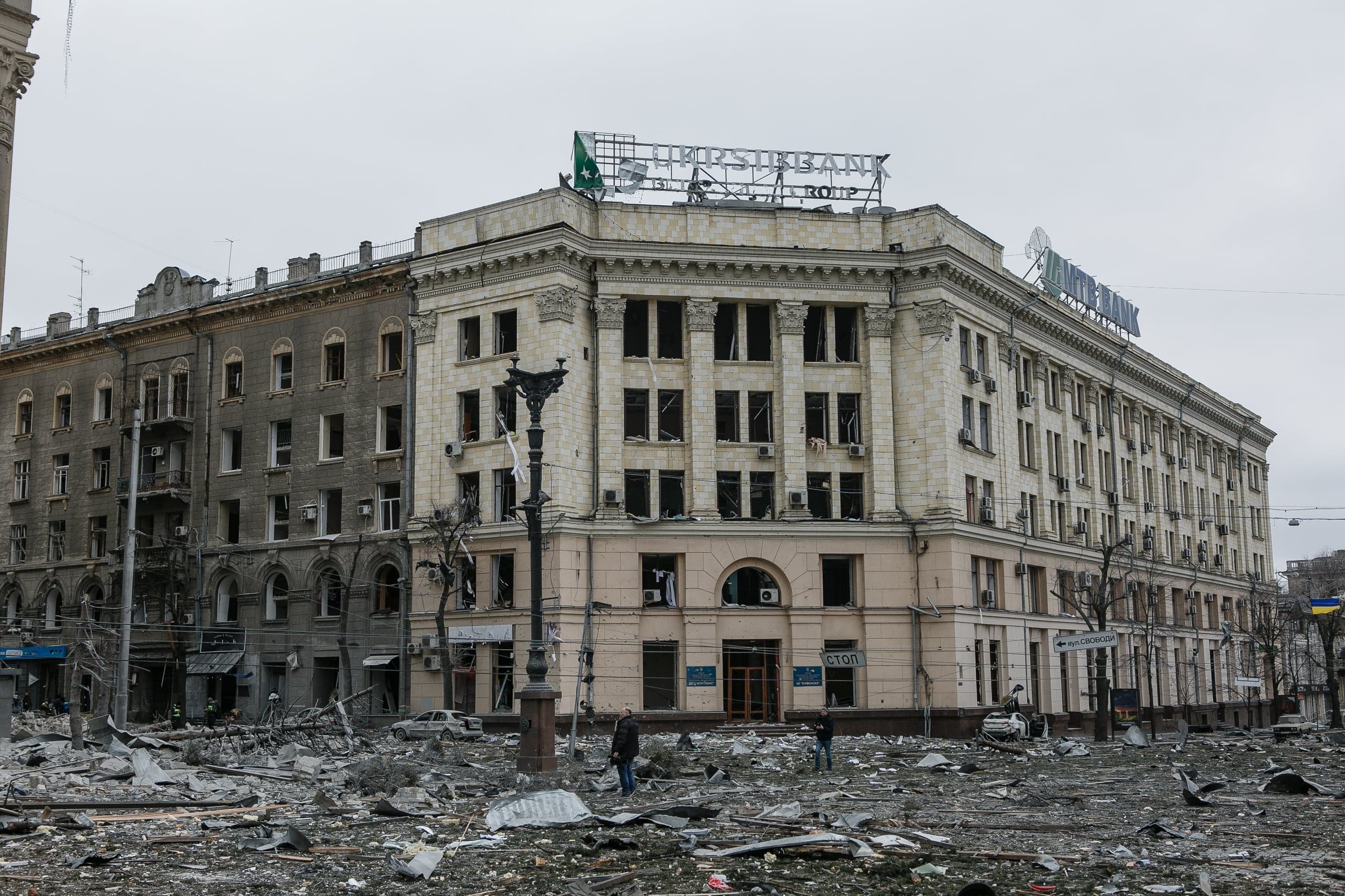 Разрушенные фасады зданий и площадь, усеянная обломками, после обстрела. На крыше виден поврежденный баннер "УКРСИББАНК".