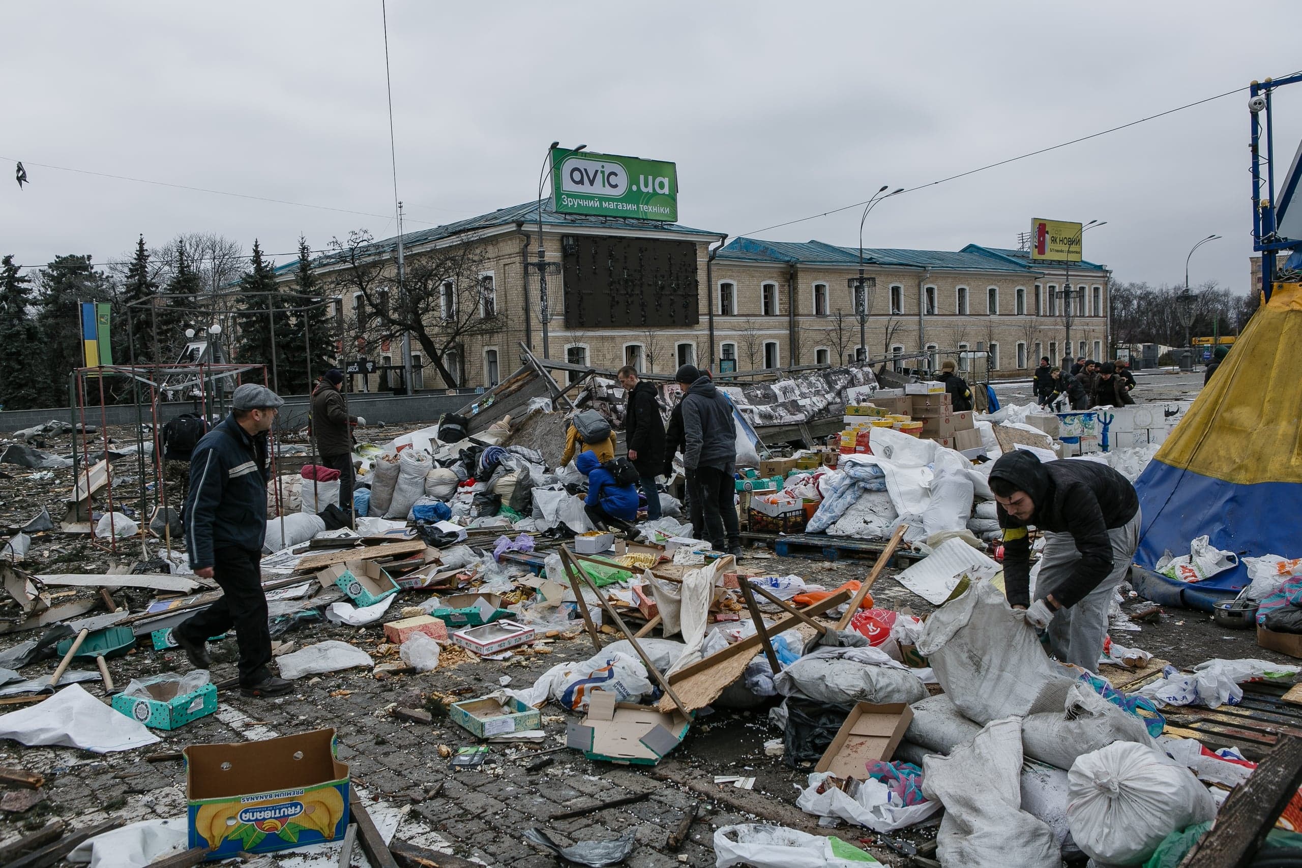 Городская площадь, заваленная обломками и мусором, где люди собирают вещи. На заднем плане здание с баннером "avic.ua Зручний магазин техніки".