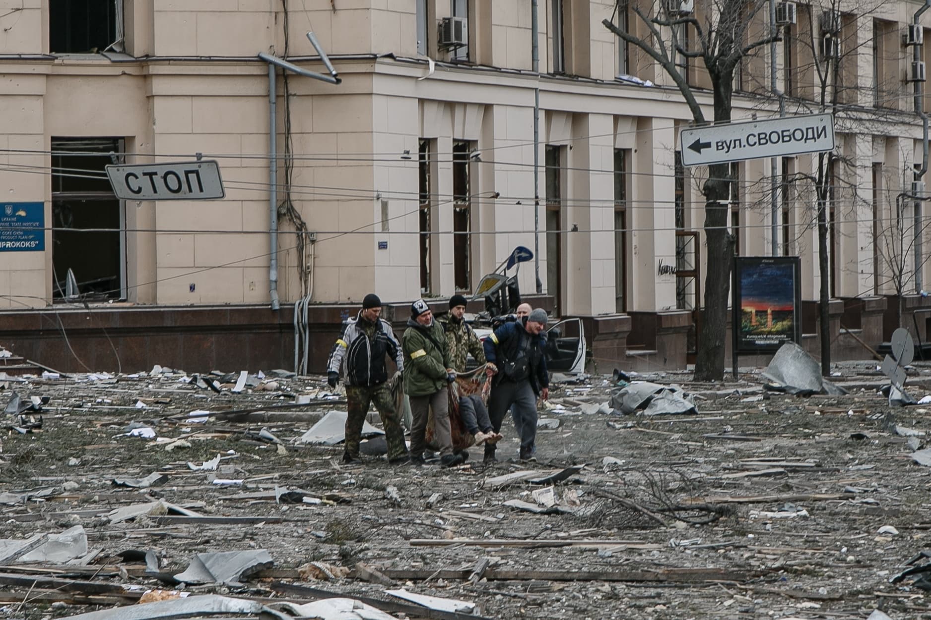 Пять человек несут тело на импровизированных носилках через заваленную обломками улицу перед разрушенным зданием. Видны дорожные знаки "СТОП" и "вул. СВОБОДИ".