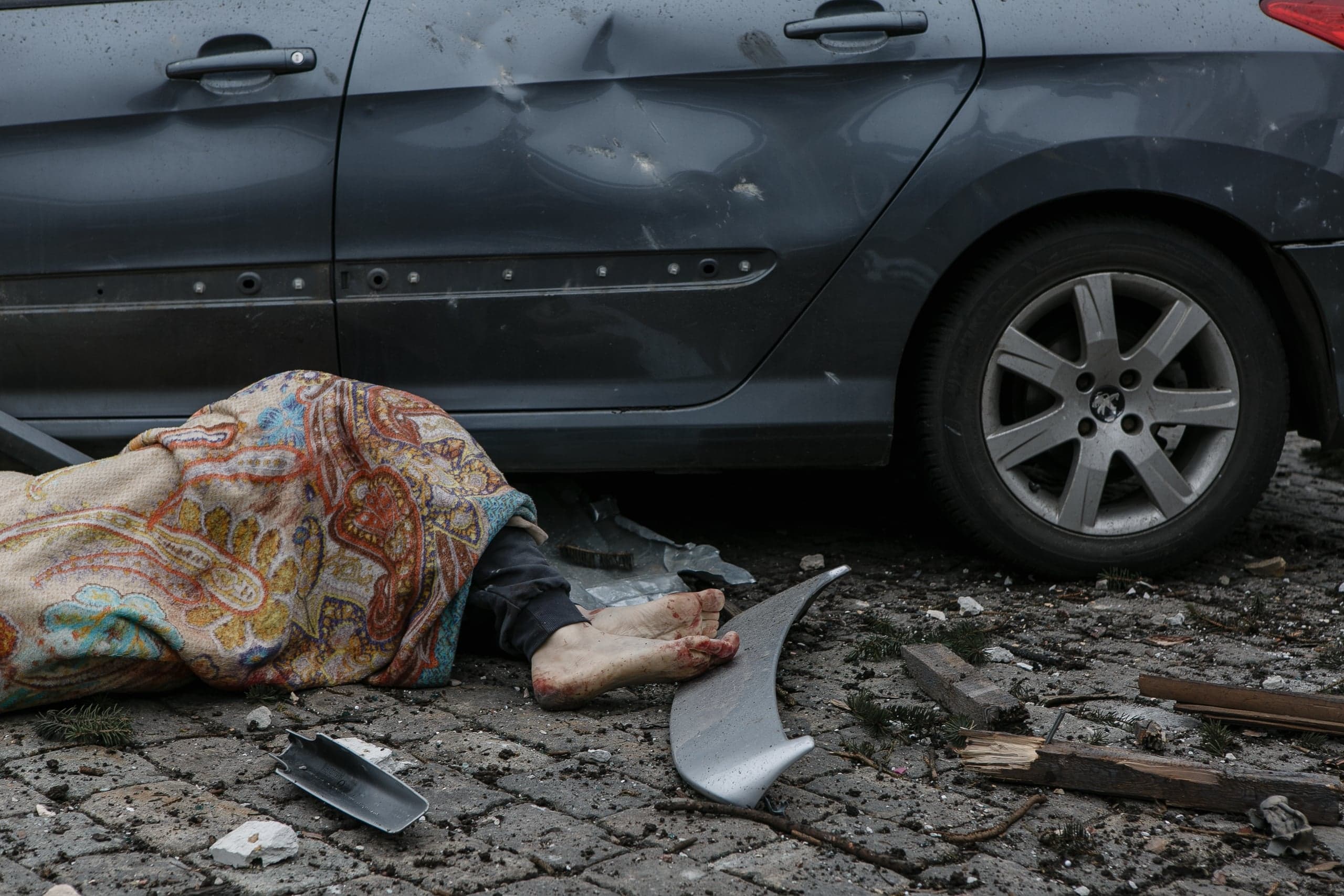 Тело, накрытое одеялом, с окровавленными ступнями лежит у поврежденного пулями или осколками серого автомобиля.