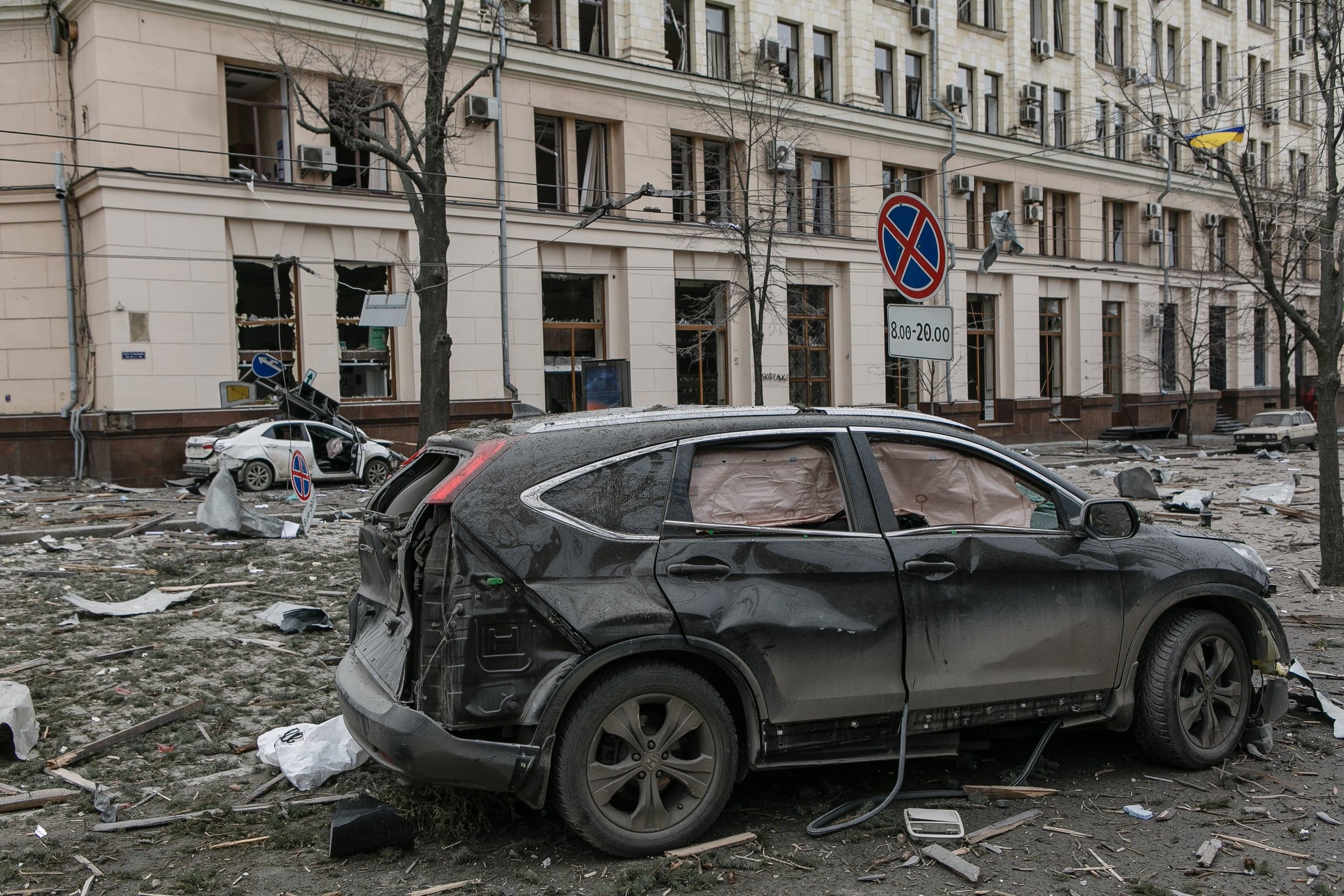 Сильно поврежденный черный автомобиль среди обломков на улице. На фоне — здание с разбитыми окнами и дорожный знак "8.00-20.00".