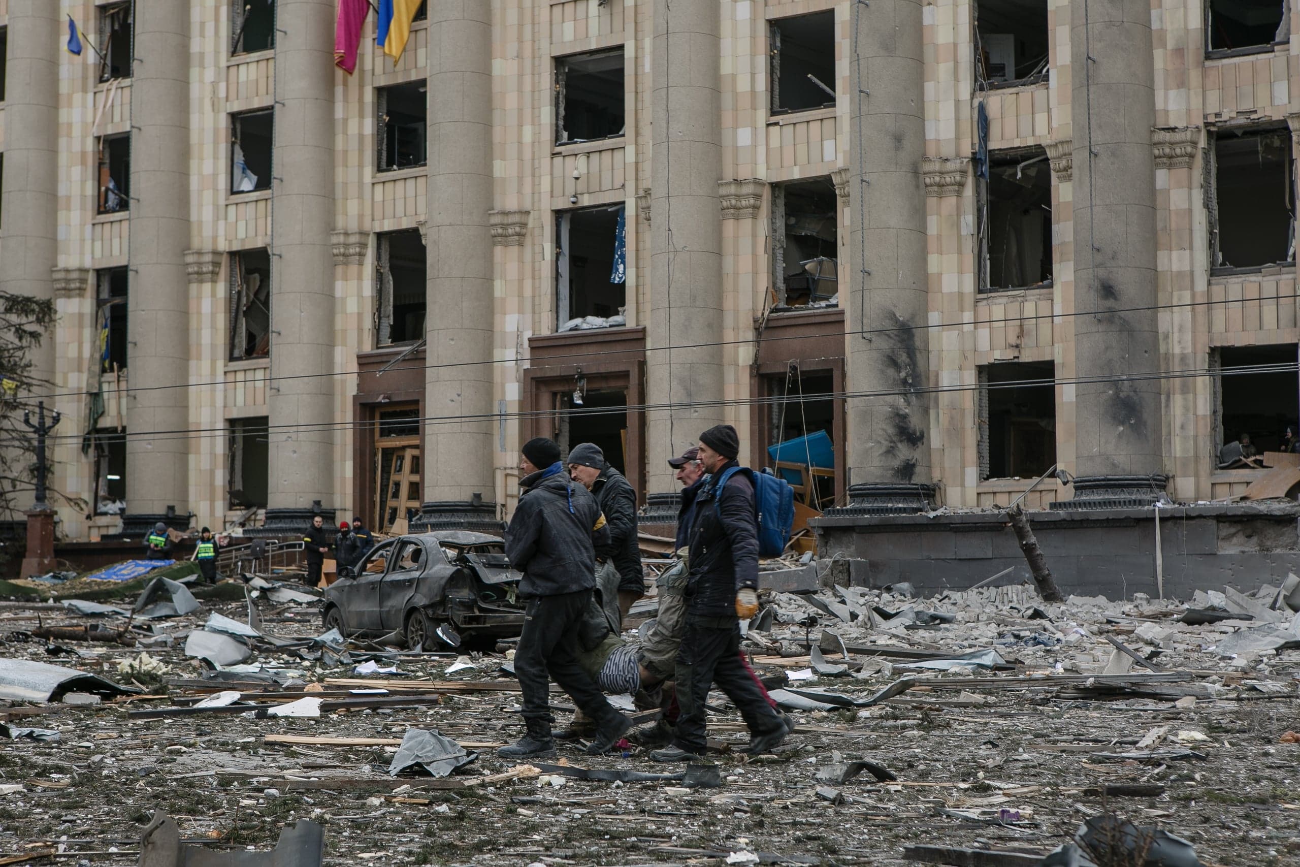 Разрушенное здание с колоннами и выбитыми окнами. На переднем плане люди идут по обломкам мимо сильно поврежденного автомобиля.
