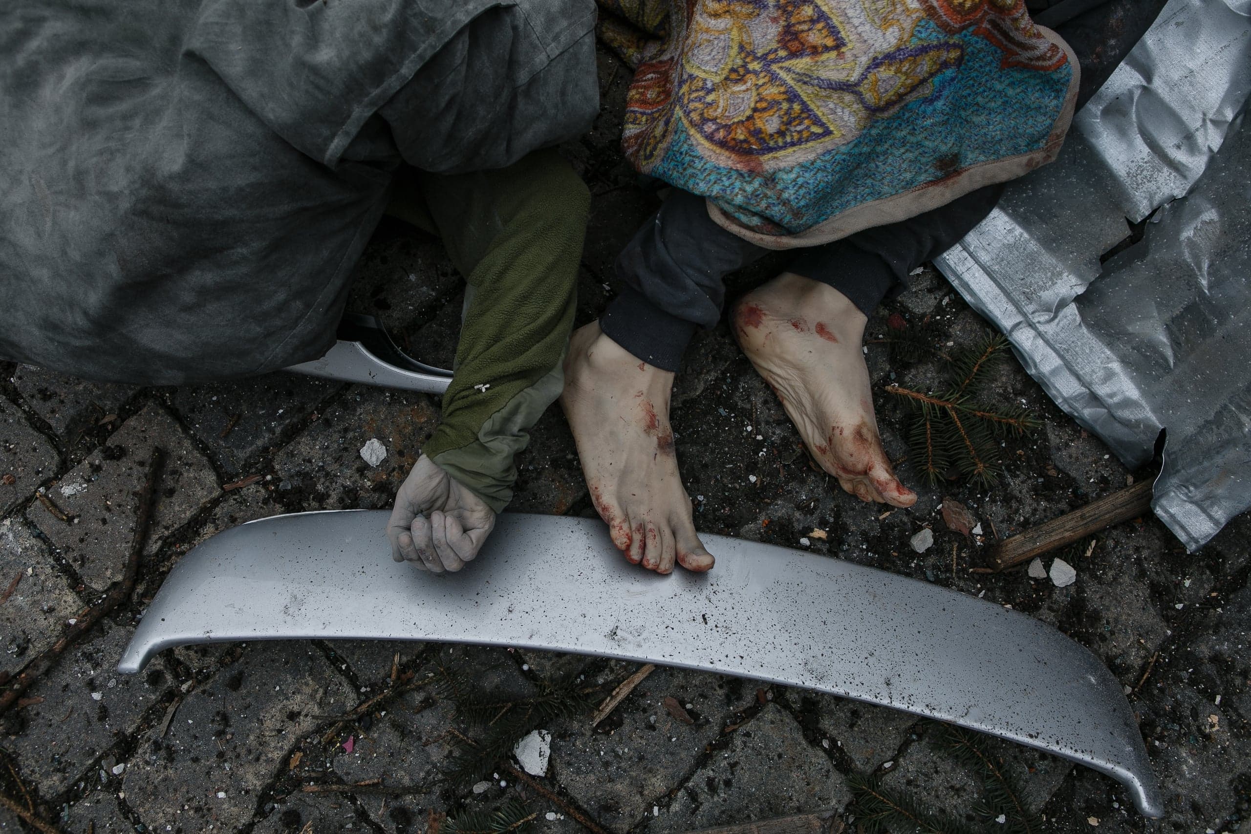 Окровавленные босые ноги и сжатый кулак двух тел среди обломков на мостовой.