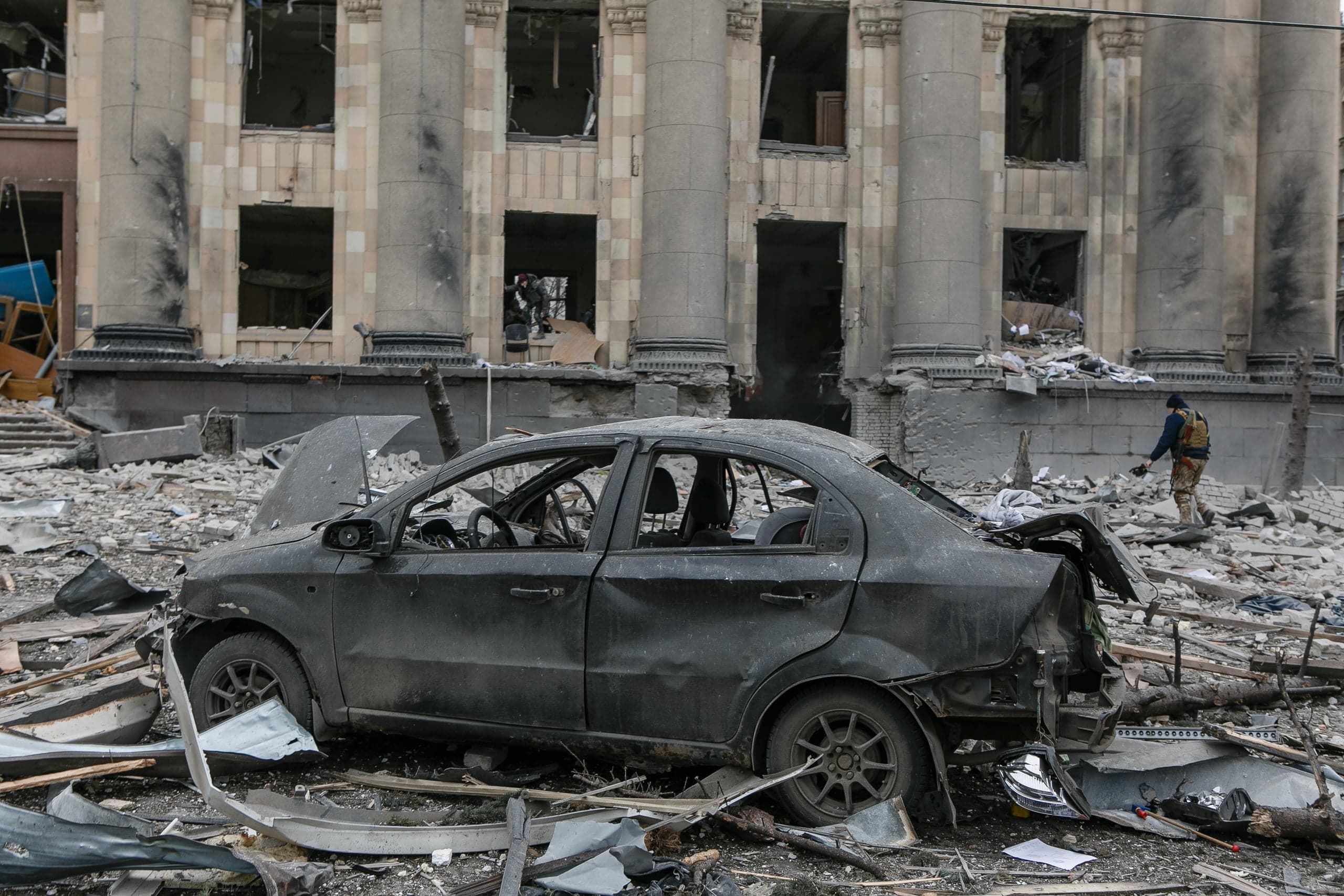 Уничтоженный автомобиль среди обломков перед сильно поврежденным зданием с колоннами. На руинах стоит человек в защитном жилете.