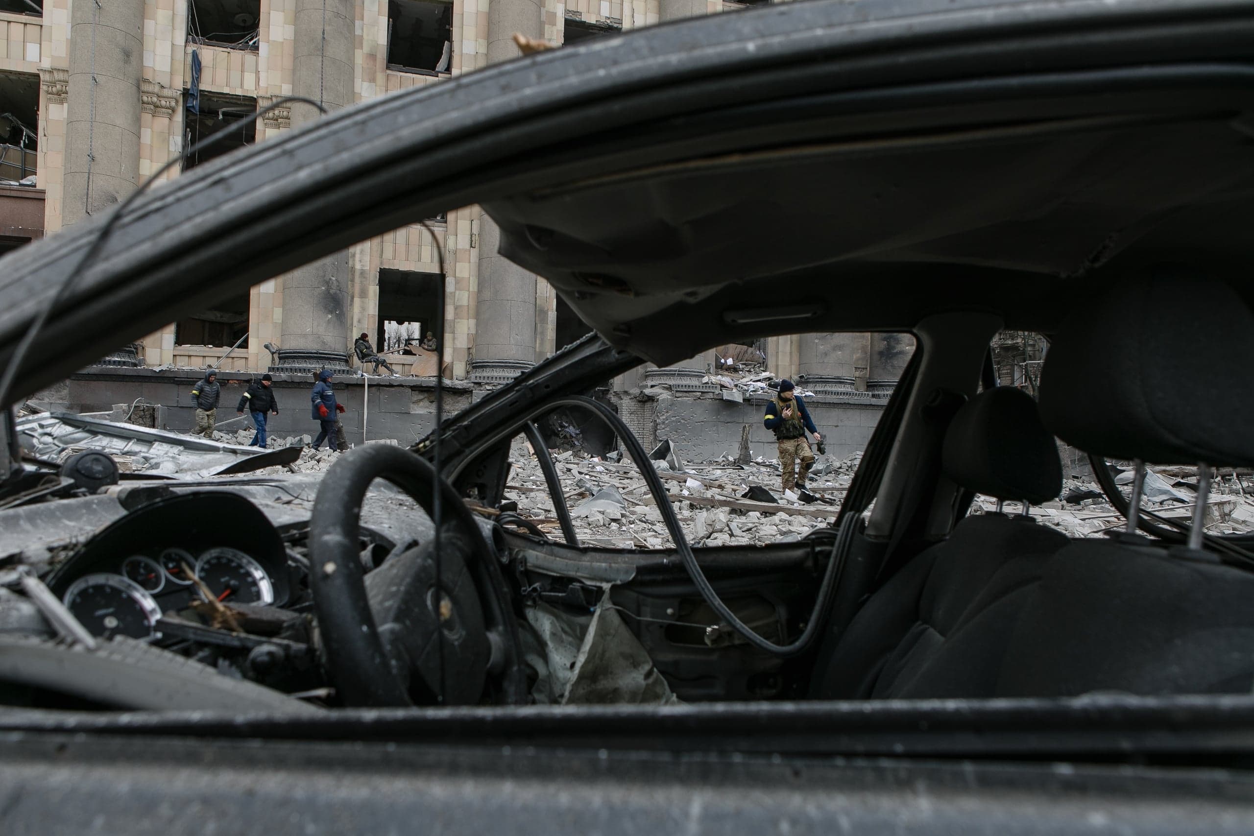 Взгляд из обгоревшего салона автомобиля на разрушенное здание с колоннами и людей, идущих по завалам.