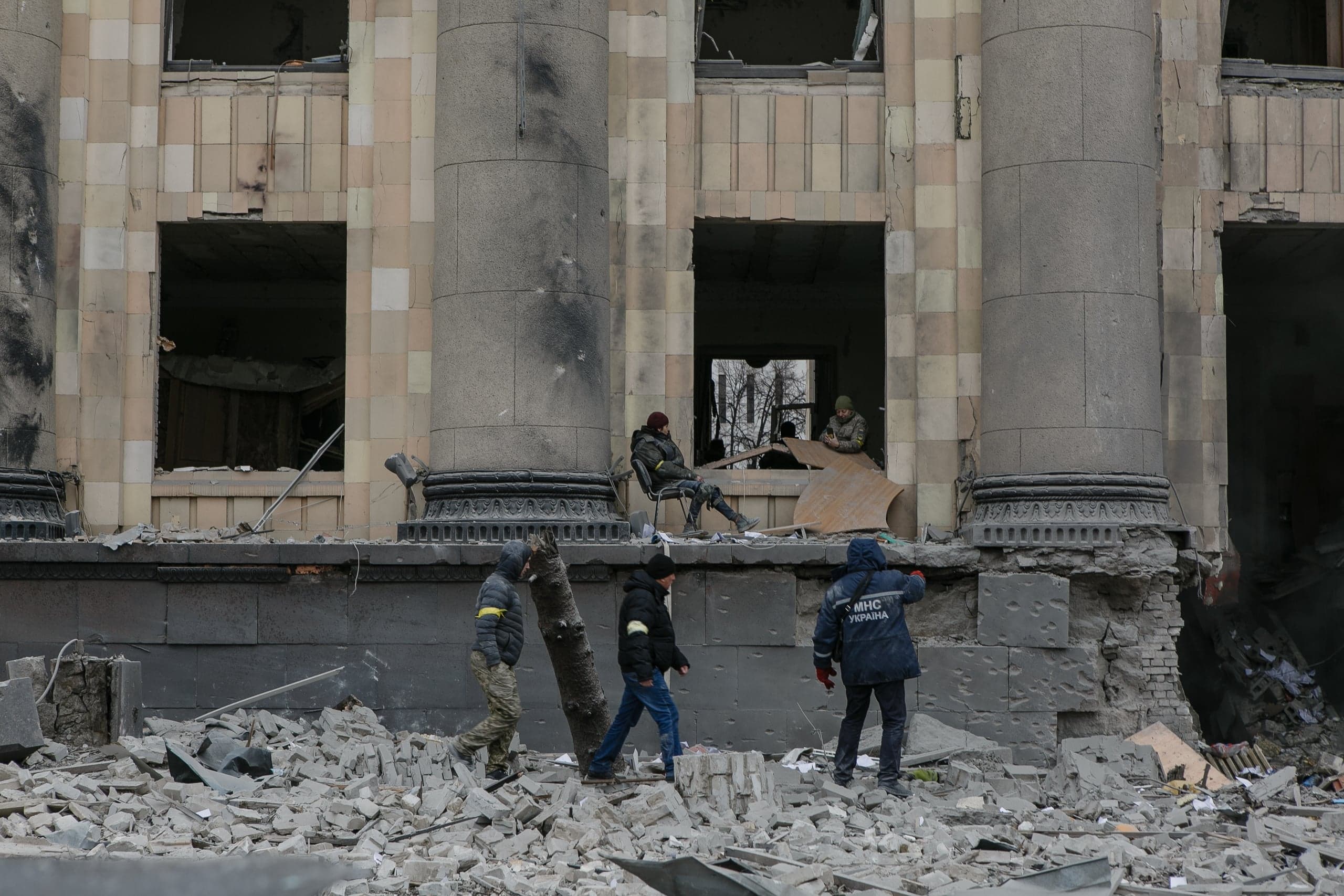 Разрушенный фасад здания с колоннами, груда обломков и люди, включая сотрудника "МНС УКРАЇНА".