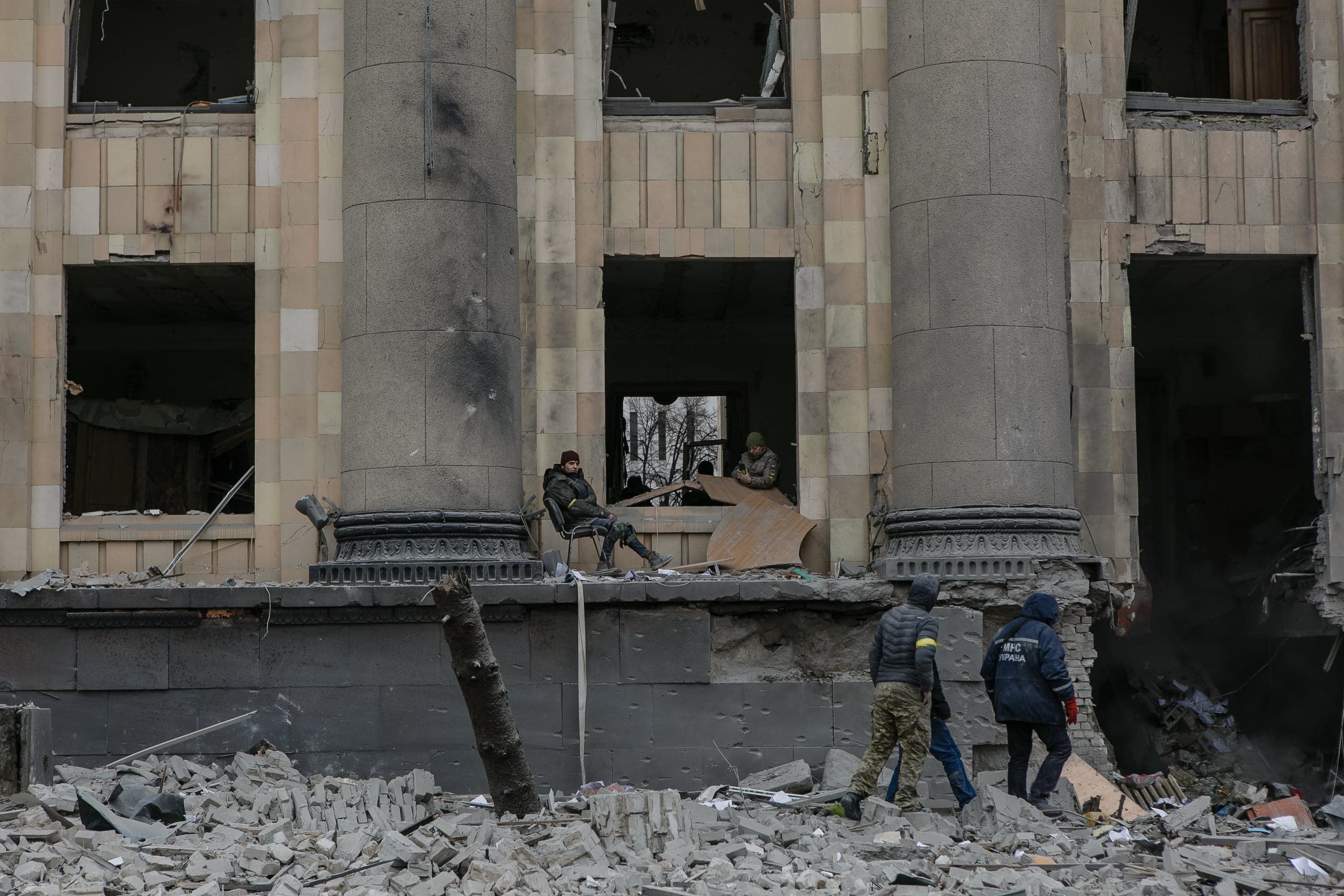 Разрушенное здание с большими колоннами, выбитыми окнами и грудой обломков на переднем плане. Два человека сидят в проеме, внизу трое мужчин (один в форме "МНС УКРАИНА") идут по руинам.