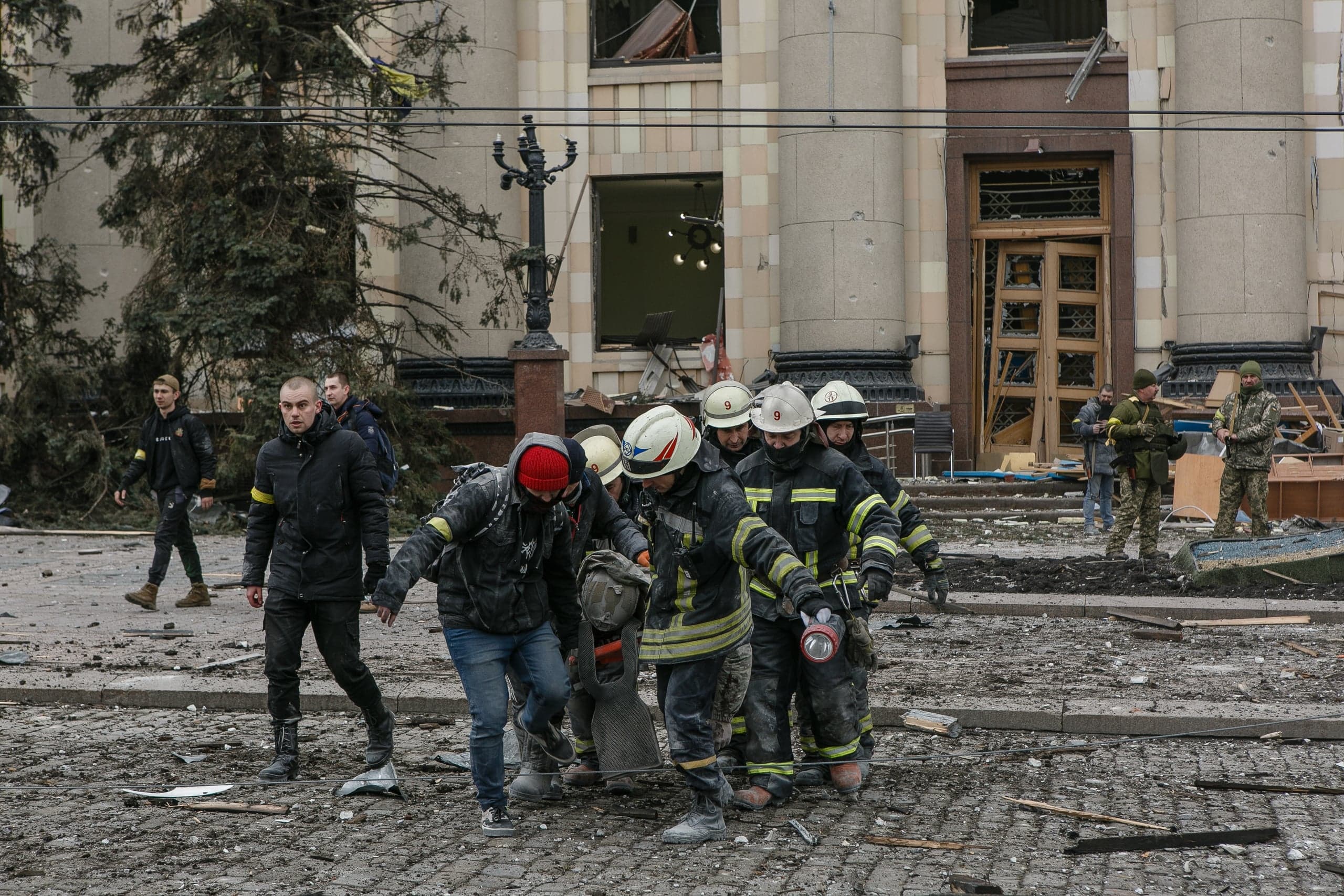 Спасатели и гражданские несут раненого мимо разрушенного фасада здания. На земле обломки, видны военные.