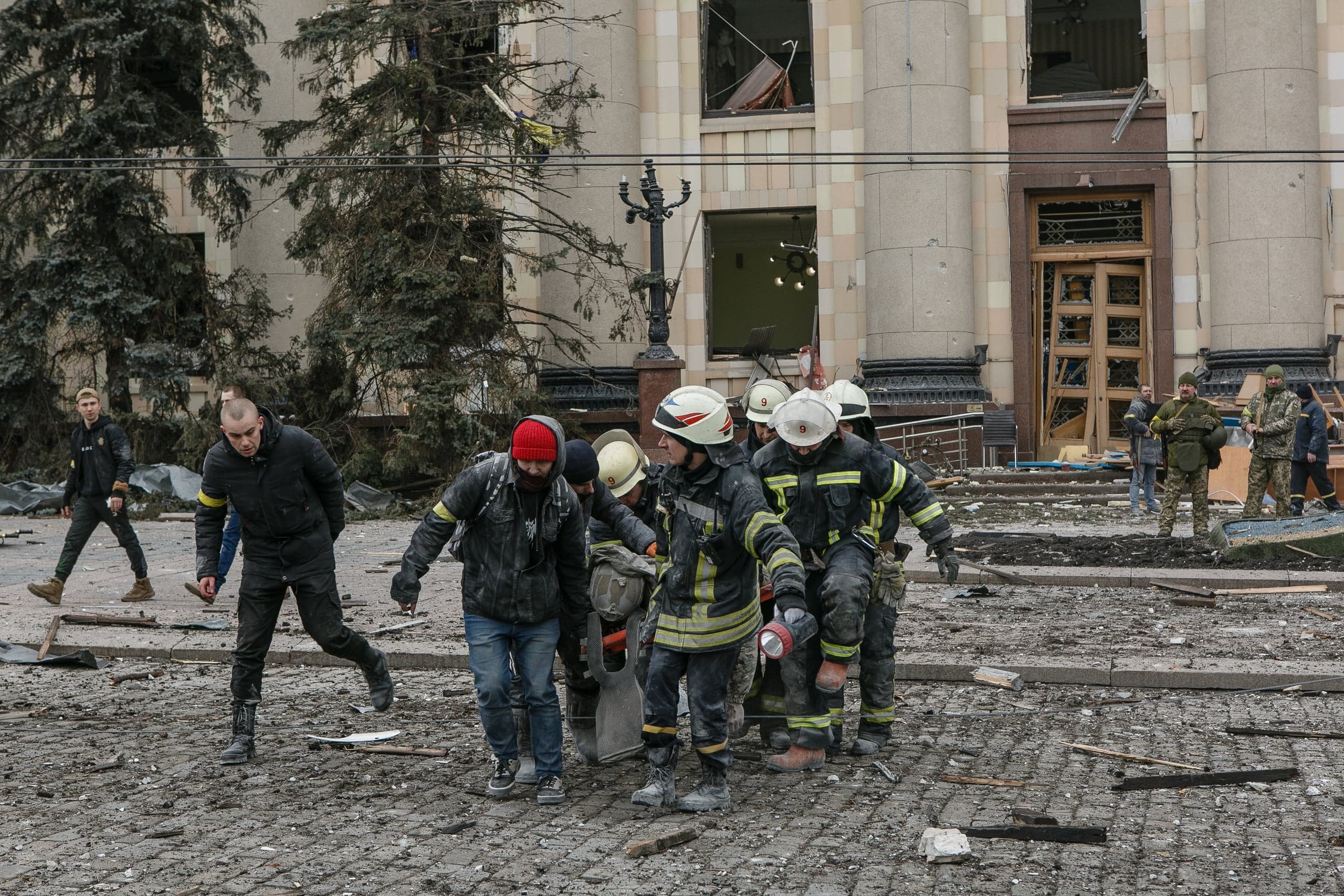 Спасатели и гражданские лица несут человека через улицу, покрытую обломками, перед сильно поврежденным зданием.