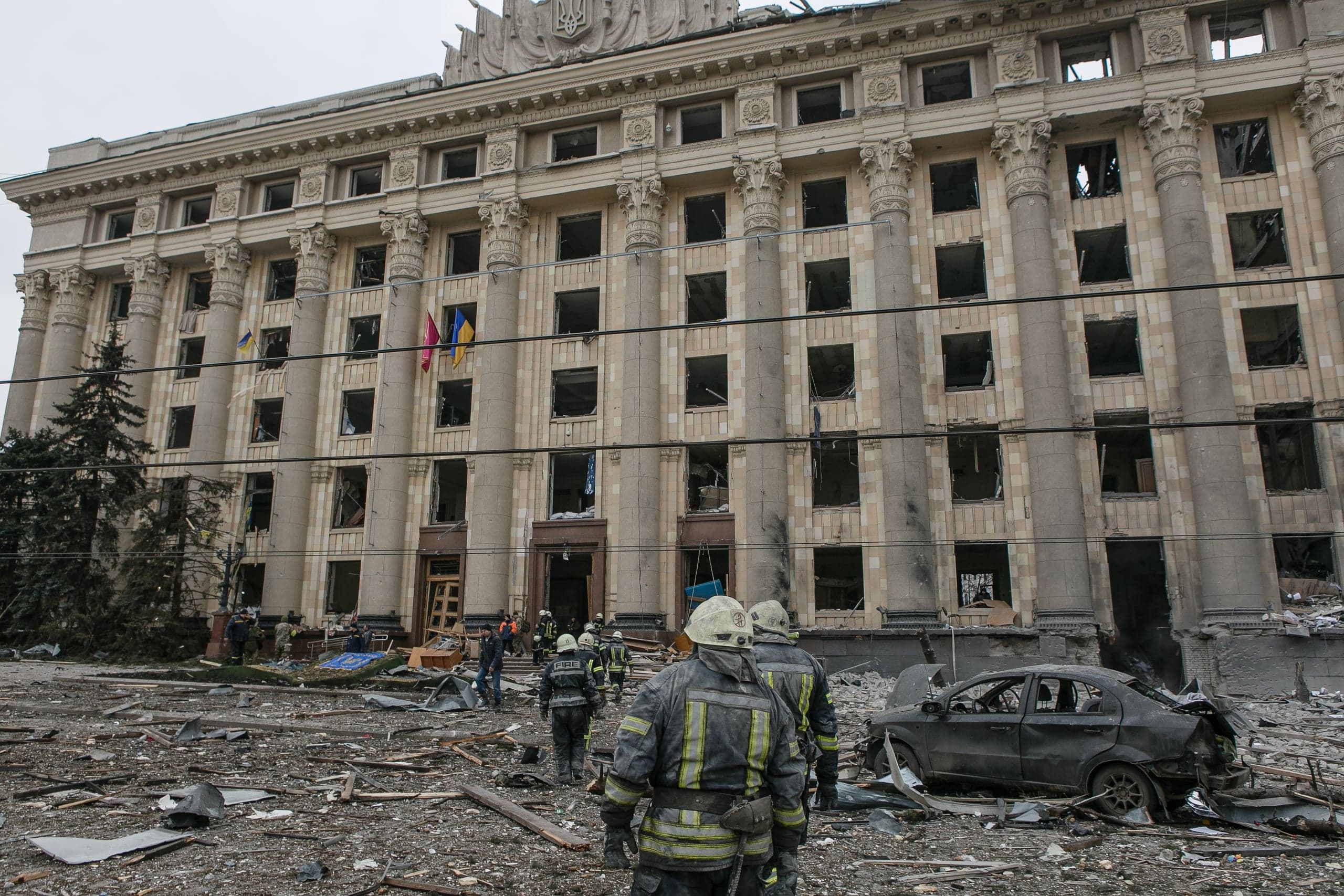 Разрушенное здание Харьковской ОГА с пустыми окнами, перед ним спасатели среди обломков и поврежденный автомобиль. Видны флаги Украины.