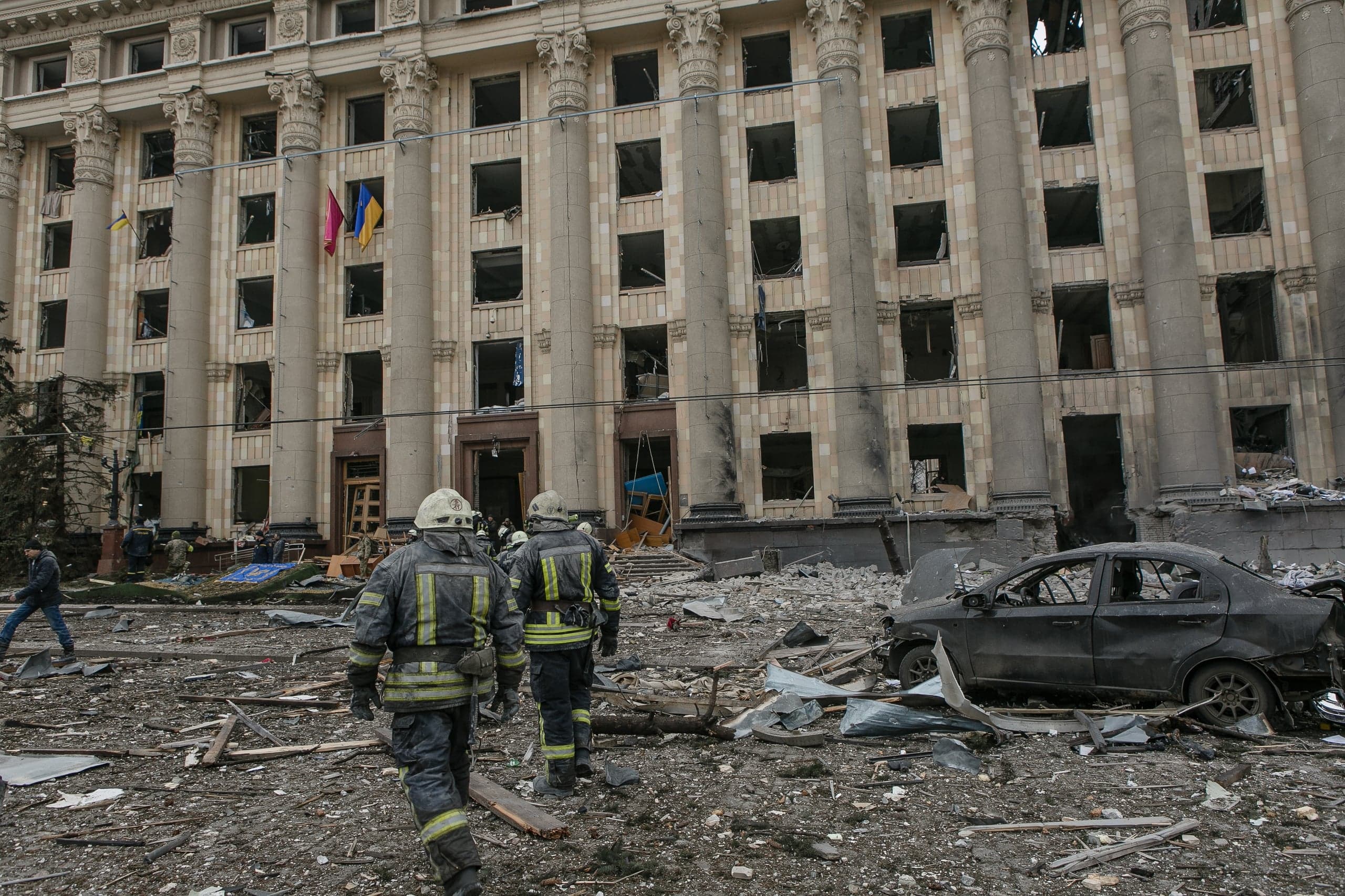 Разрушенное здание с колоннами, выбитыми окнами и флагами Украины. На переднем плане — два спасателя и обломки, включая сгоревший автомобиль.