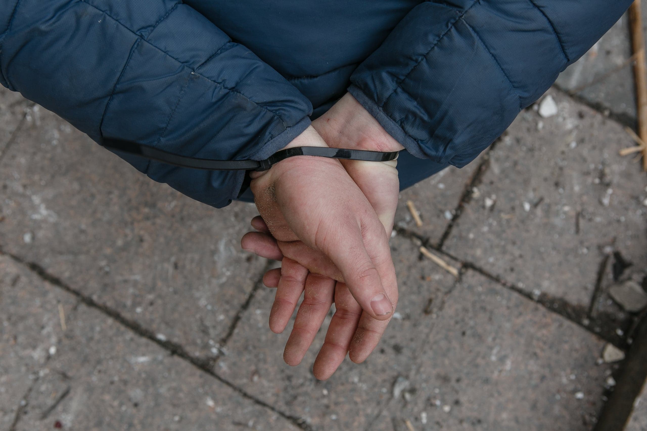Связанные пластиковым хомутом руки человека в синей куртке, стоящего на бетонной плитке.