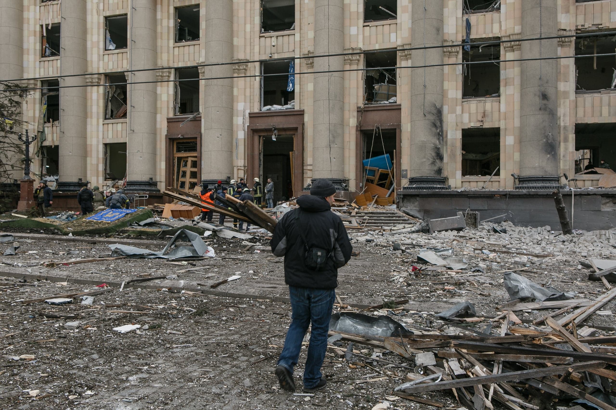 Разрушенный фасад здания с колоннами и площадь, заваленная обломками. Люди расчищают завалы.