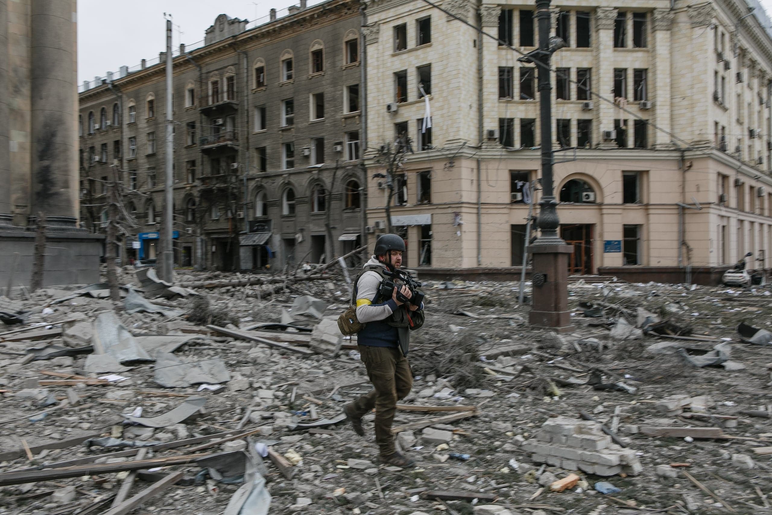 Журналист в шлеме и бронежилете бежит по площади, полностью покрытой обломками и строительным мусором. Здания на фоне разрушены и имеют выбитые окна.
