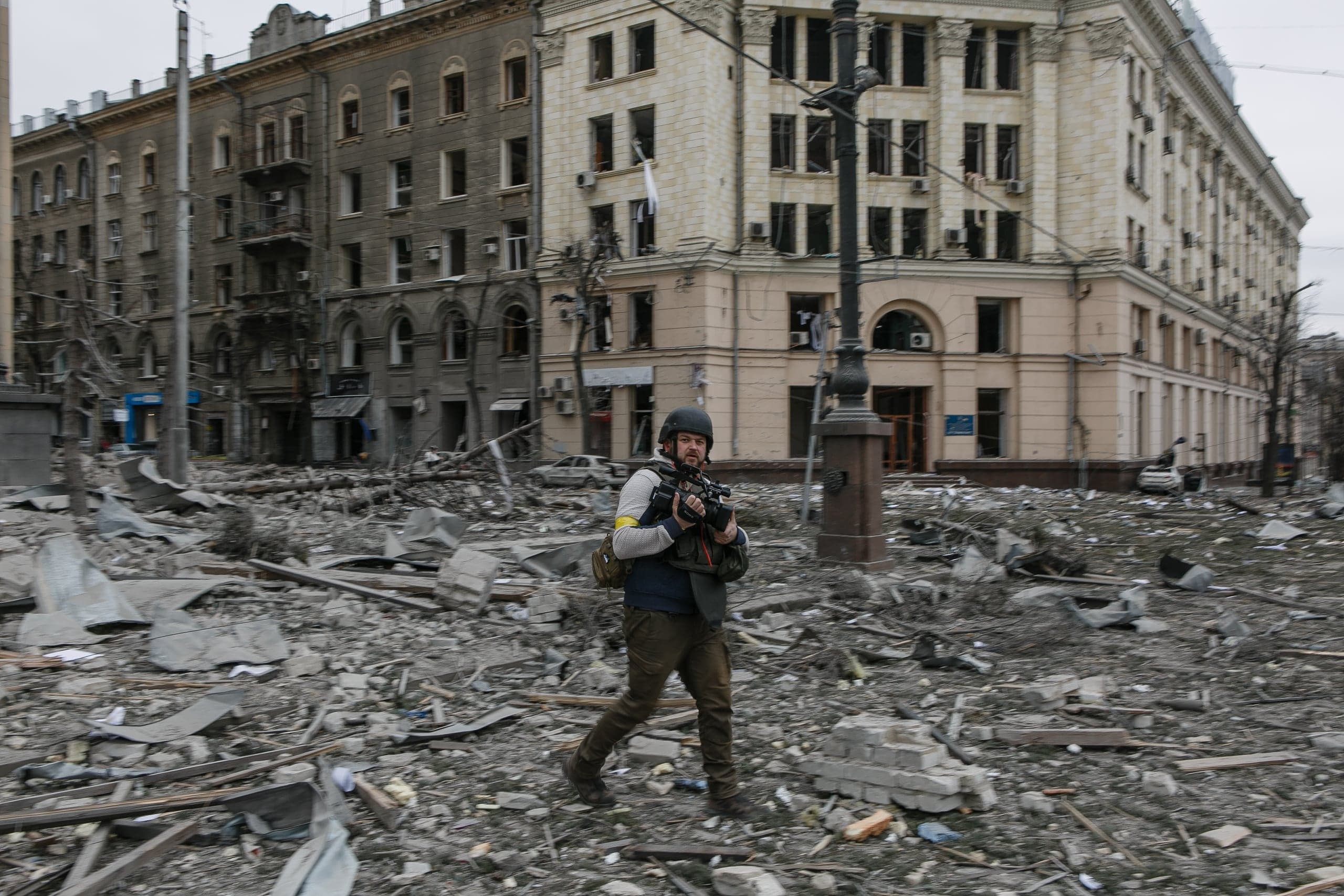 Журналист в каске и бронежилете идет через площадь, заваленную обломками и мусором, на фоне сильно поврежденных зданий.