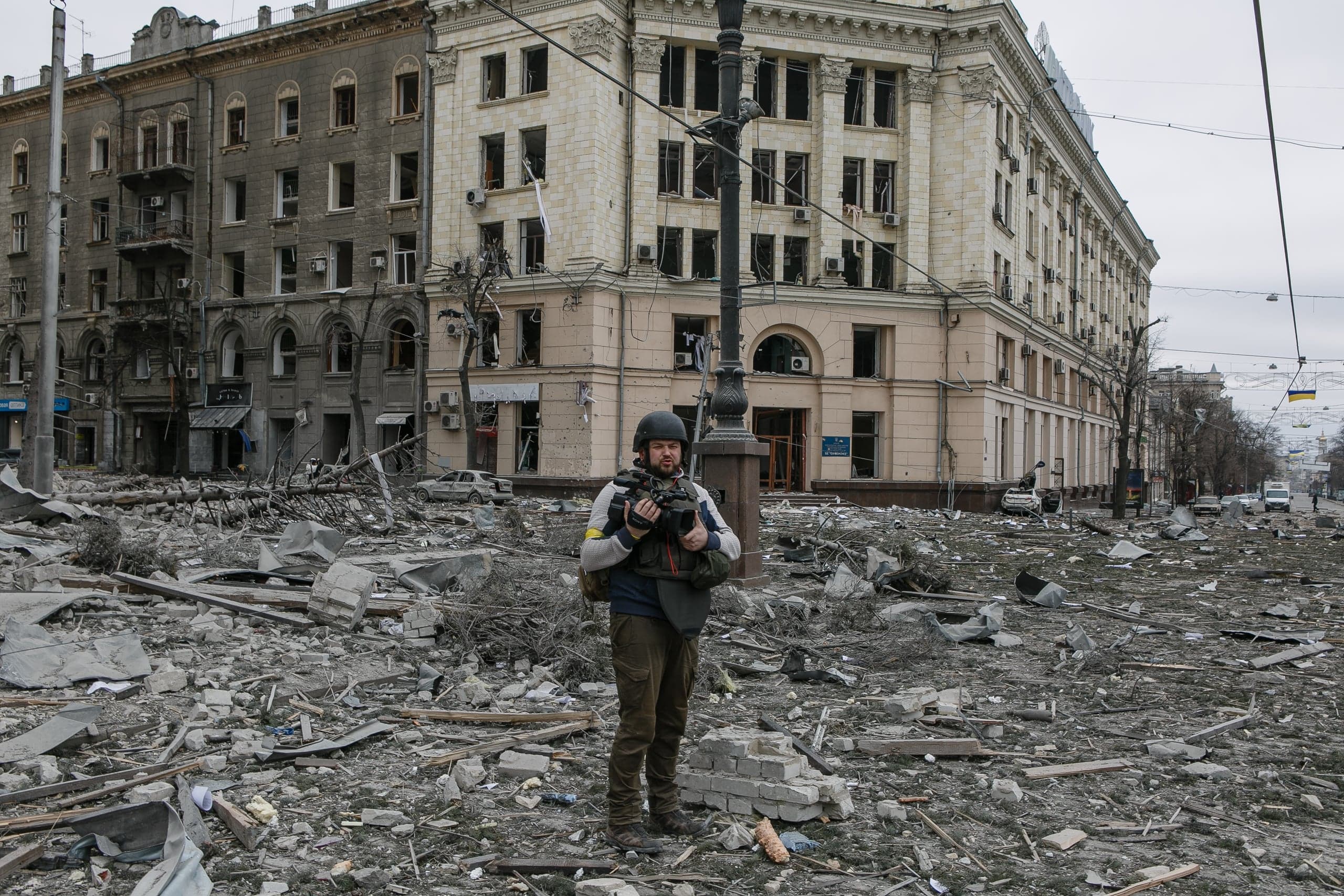 Журналист в защитной экипировке и шлеме стоит среди обширных руин и обломков. Позади видны сильно поврежденные взрывом городские здания.