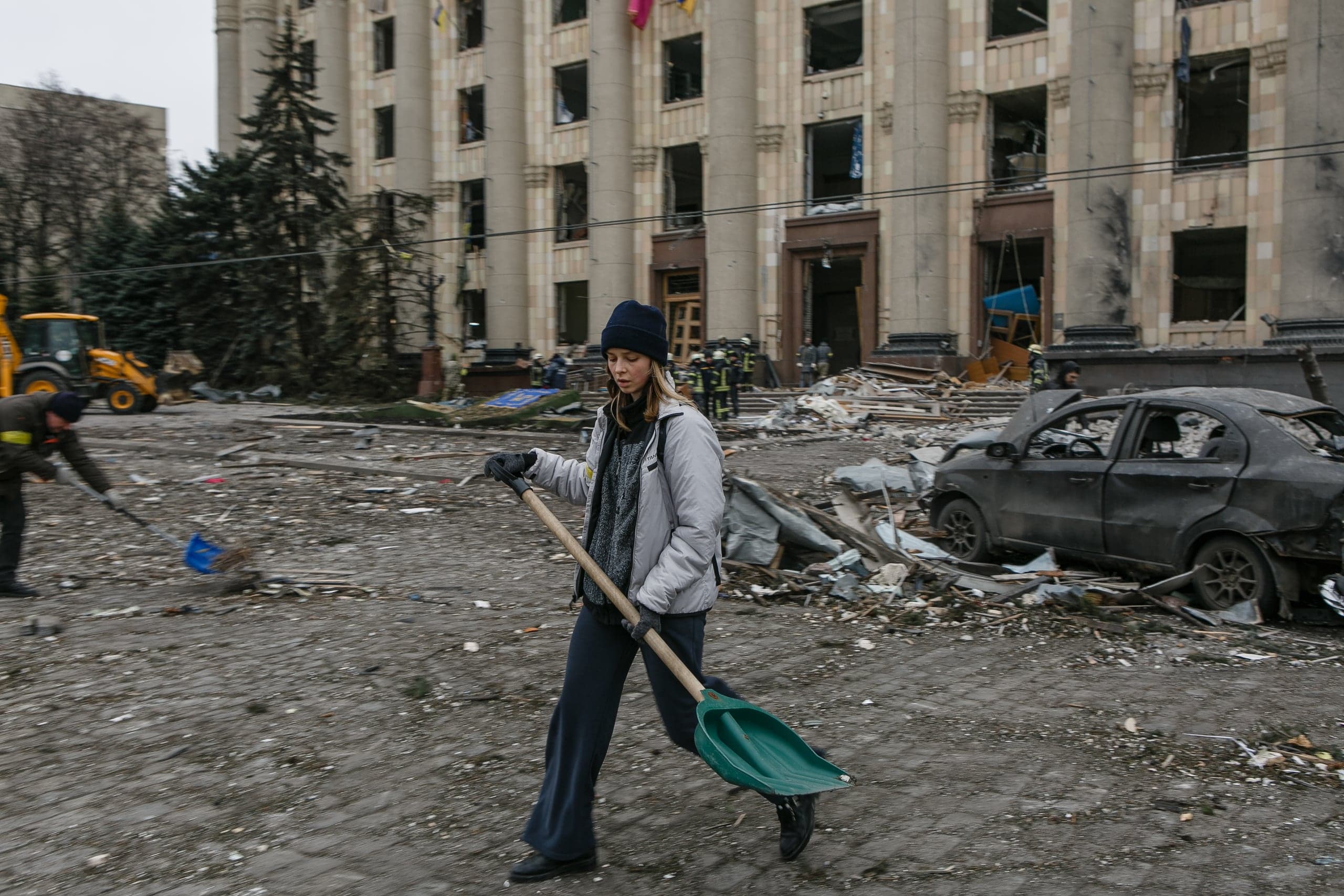 Женщина с зеленой лопатой убирает обломки перед сильно поврежденным зданием с колоннами; на земле — груды мусора и остов сгоревшего автомобиля.