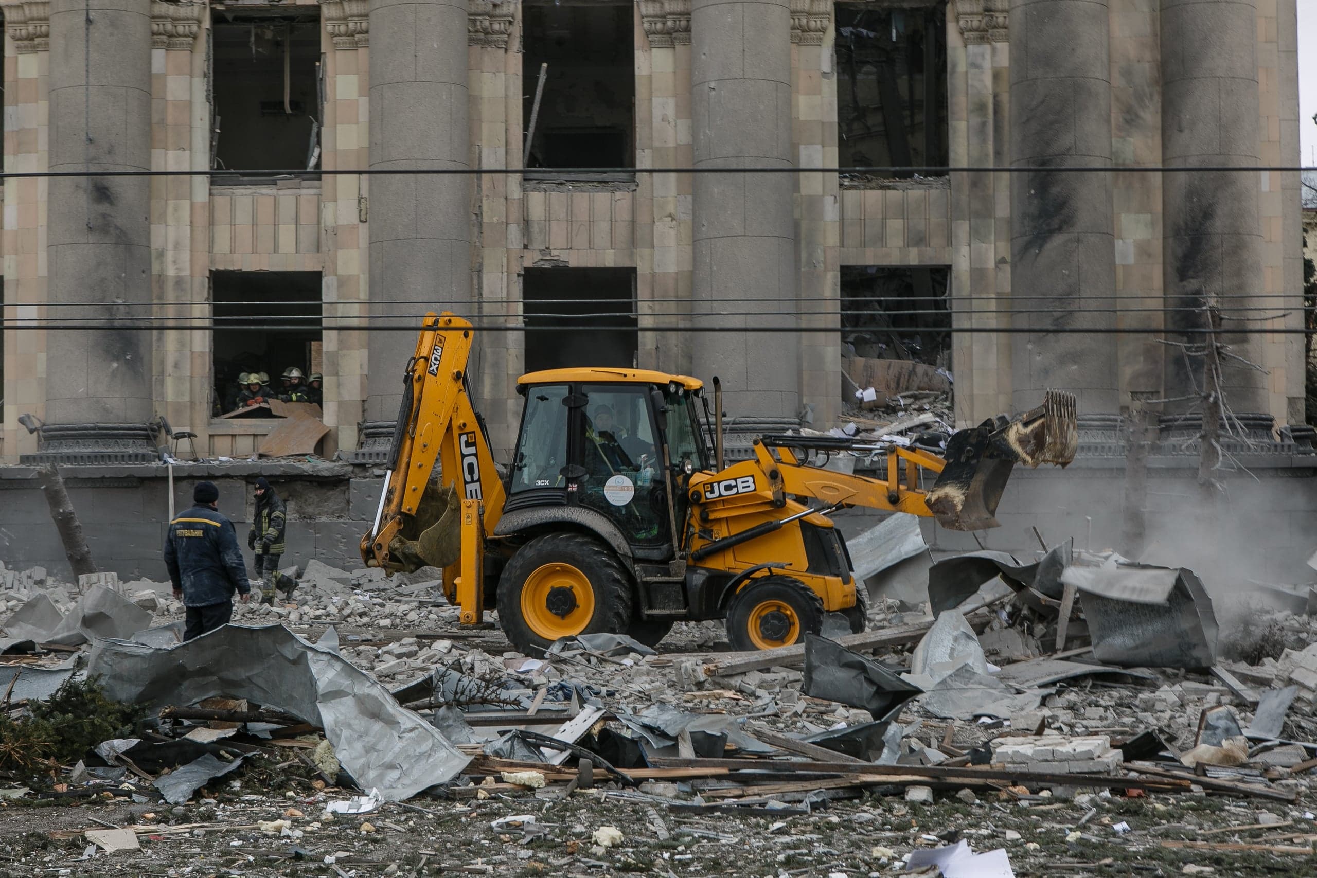 Желтый экскаватор JCB расчищает завалы на фоне сильно поврежденного монументального здания с колоннами. На переднем плане обломки и два человека в рабочей одежде.