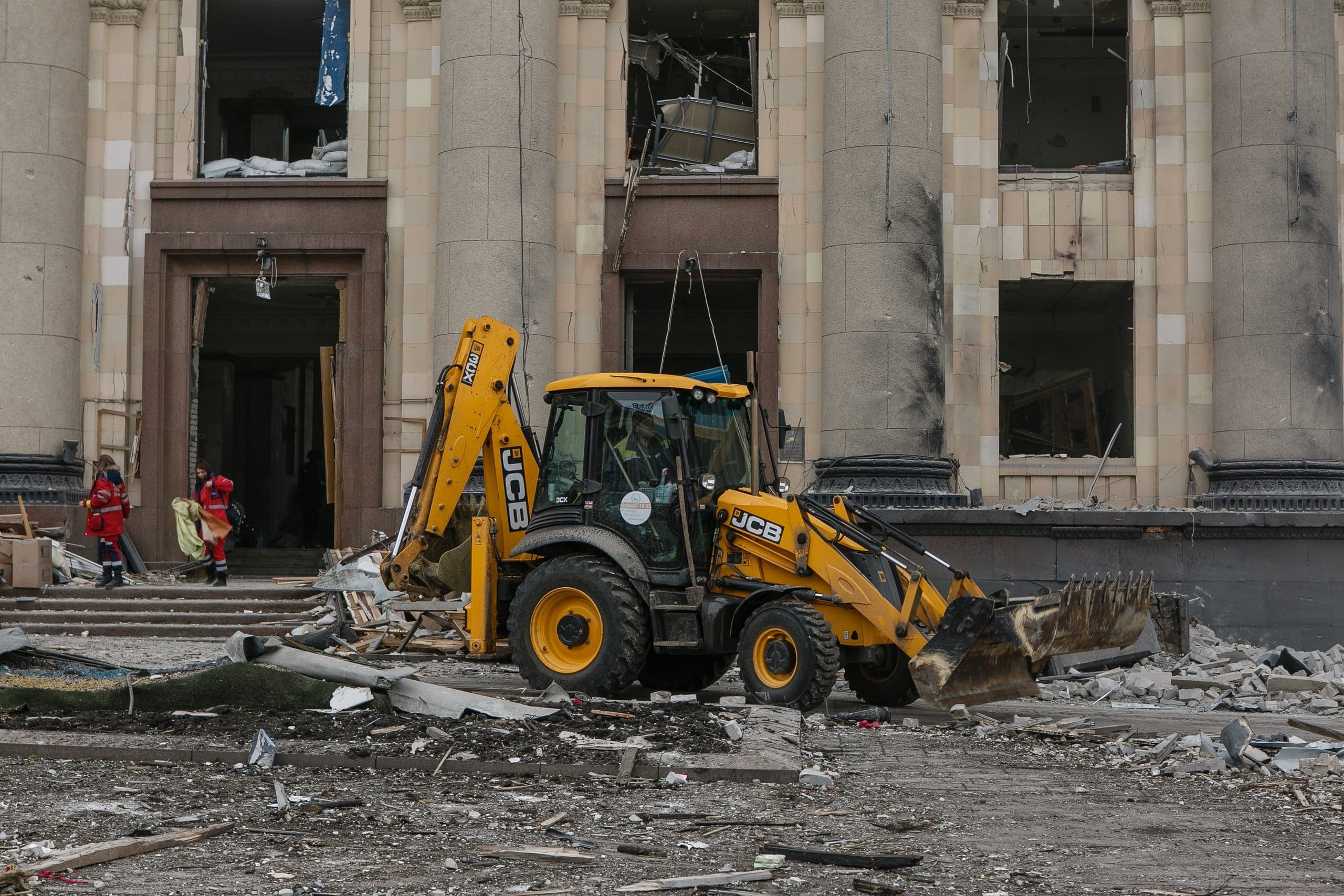 Желтый экскаватор-погрузчик JCB на фоне сильно поврежденного обстрелом здания с колоннами. На земле много строительных обломков.