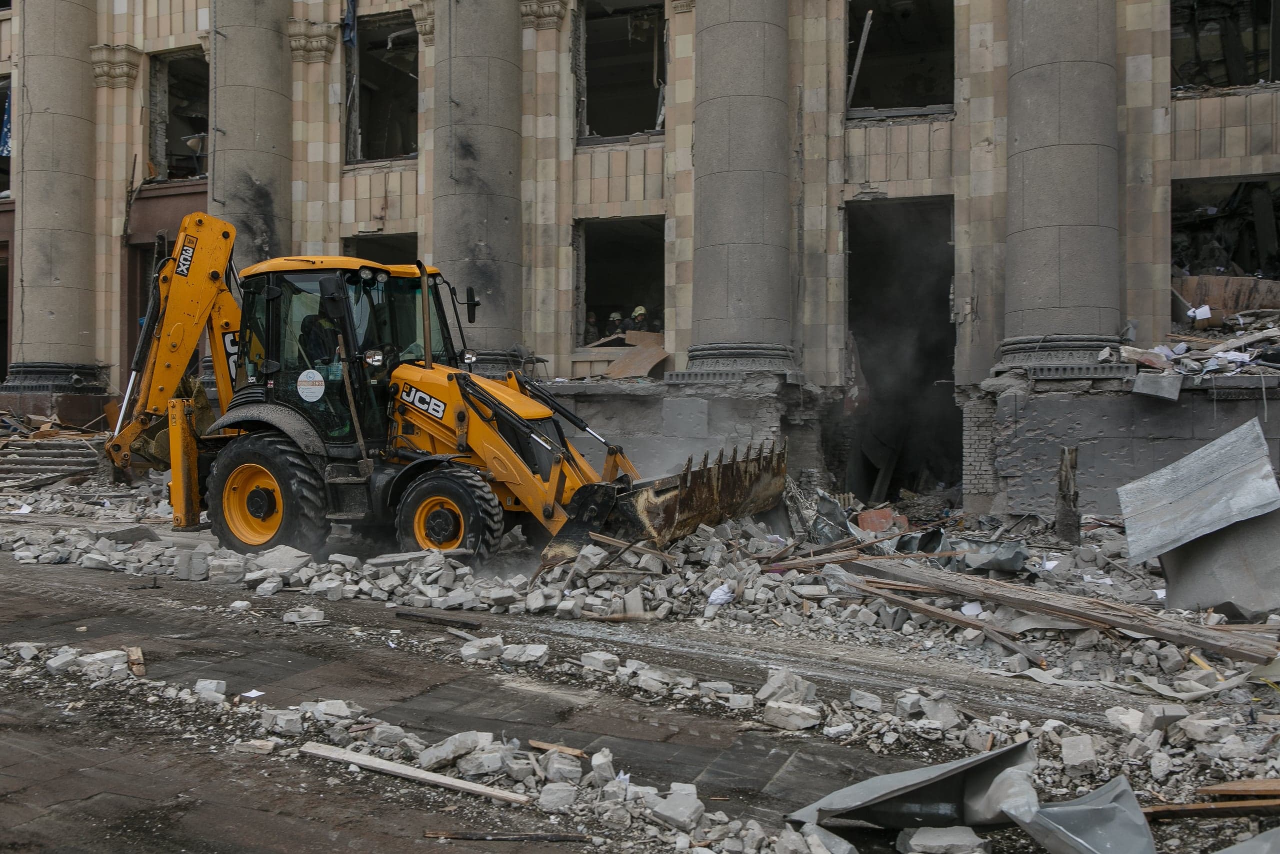 Желтый экскаватор-погрузчик JCB убирает строительный мусор на фоне разрушенного здания с колоннами.