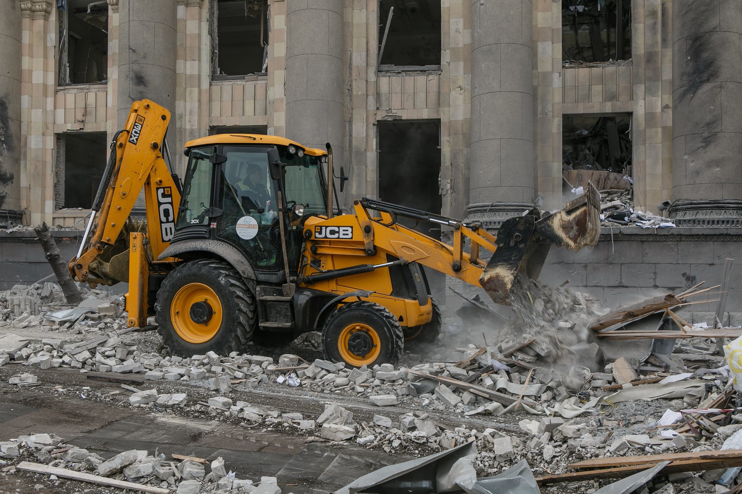 Желтый экскаватор "JCB" убирает груды обломков на фоне разрушенного здания с колоннами.