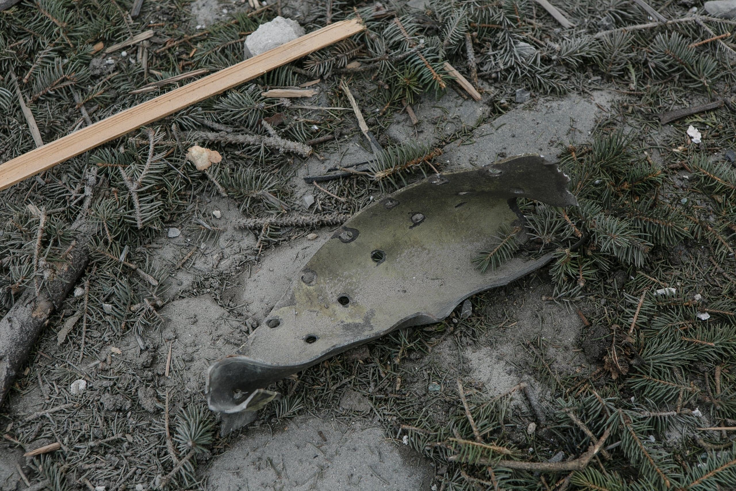 Фрагмент металла с отверстиями (вероятно, обшивка ракеты/дрона) лежит на земле среди песка и хвойных веток.