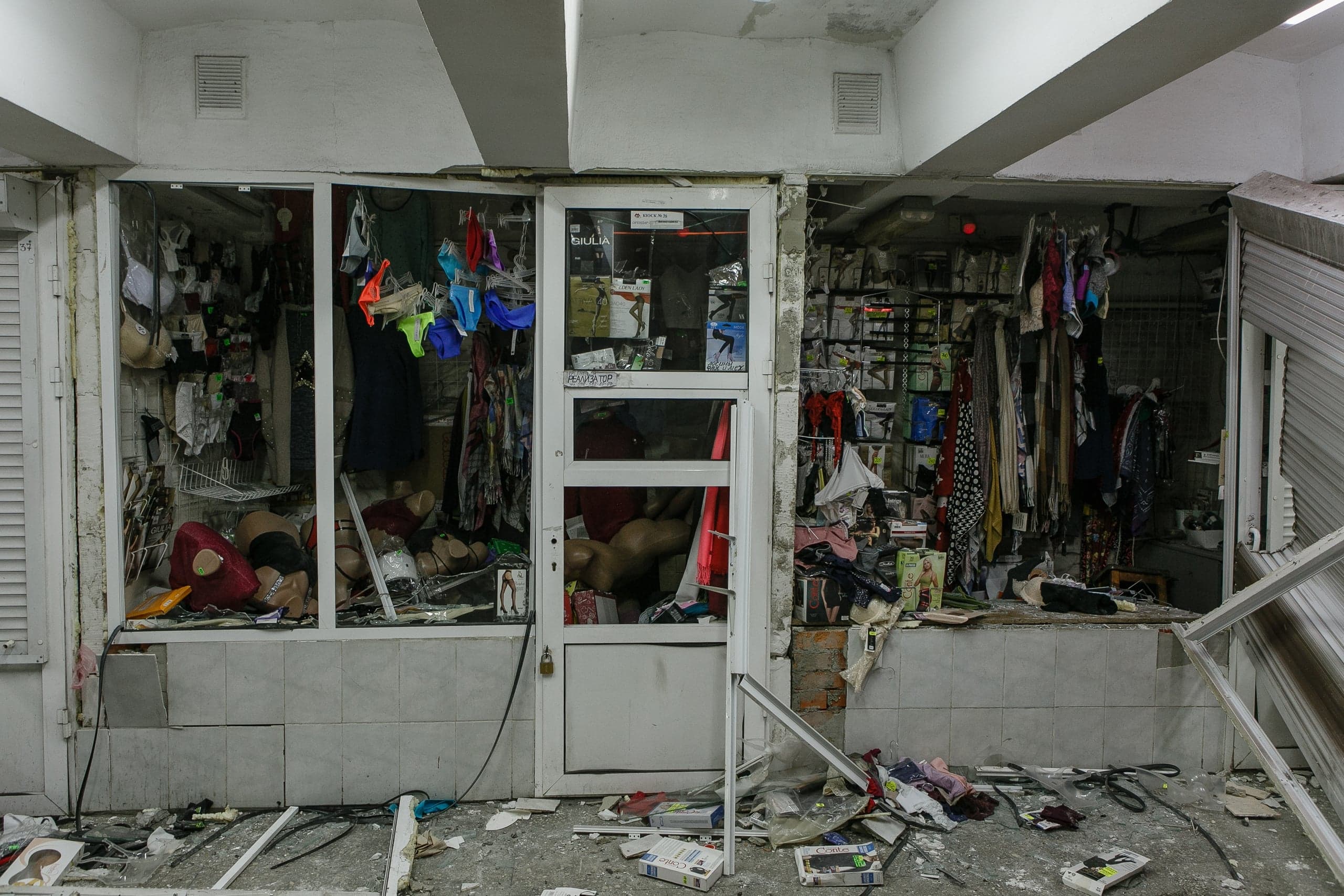 Сильно поврежденный фасад магазина нижнего белья с разбитыми окнами, разбросанными товарами и деталями манекенов, искореженными рольставнями.