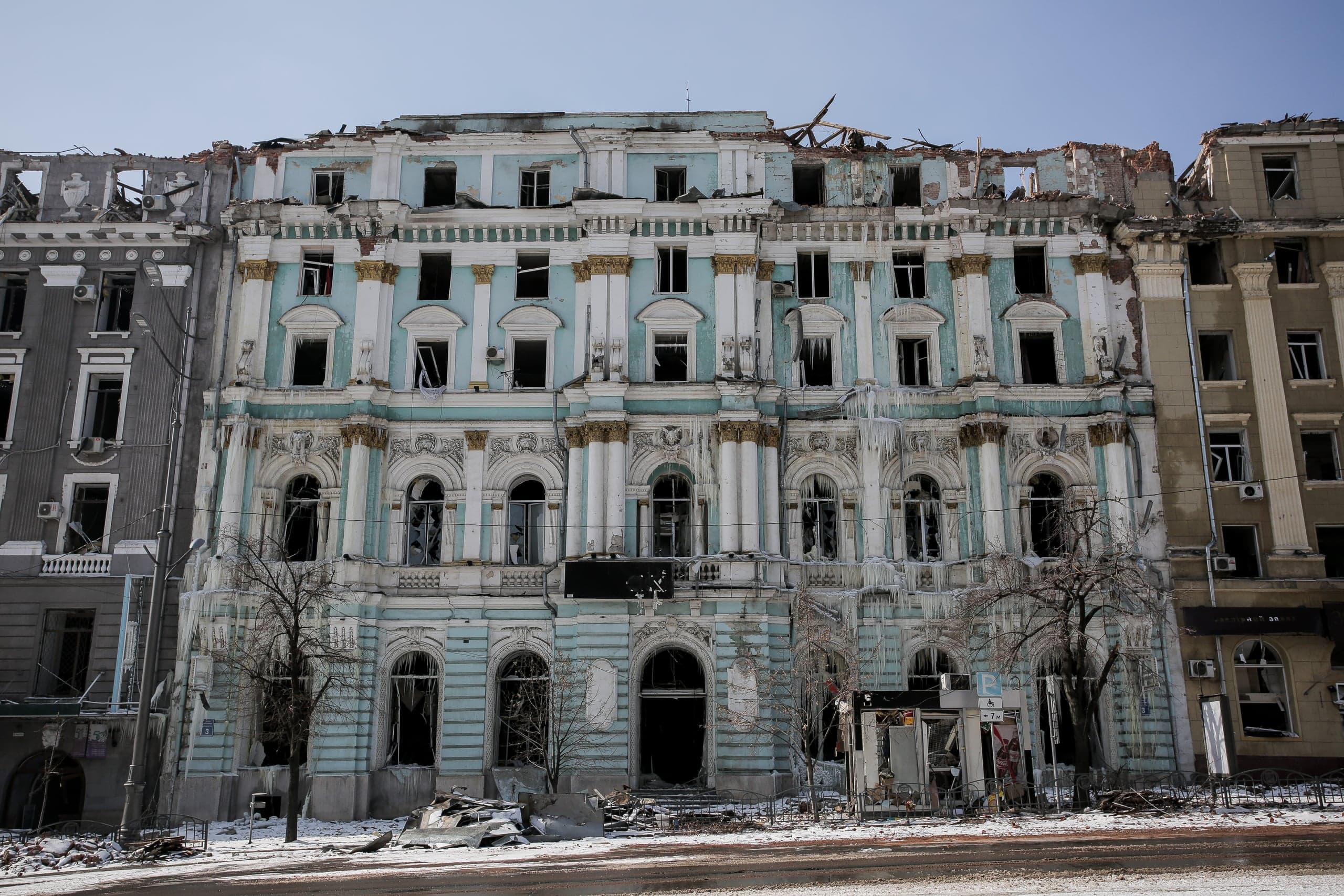 Разрушенное бирюзовое здание с выбитыми окнами, поврежденной крышей и сосульками на фасаде, стоящее посреди улицы, покрытой снегом и обломками.