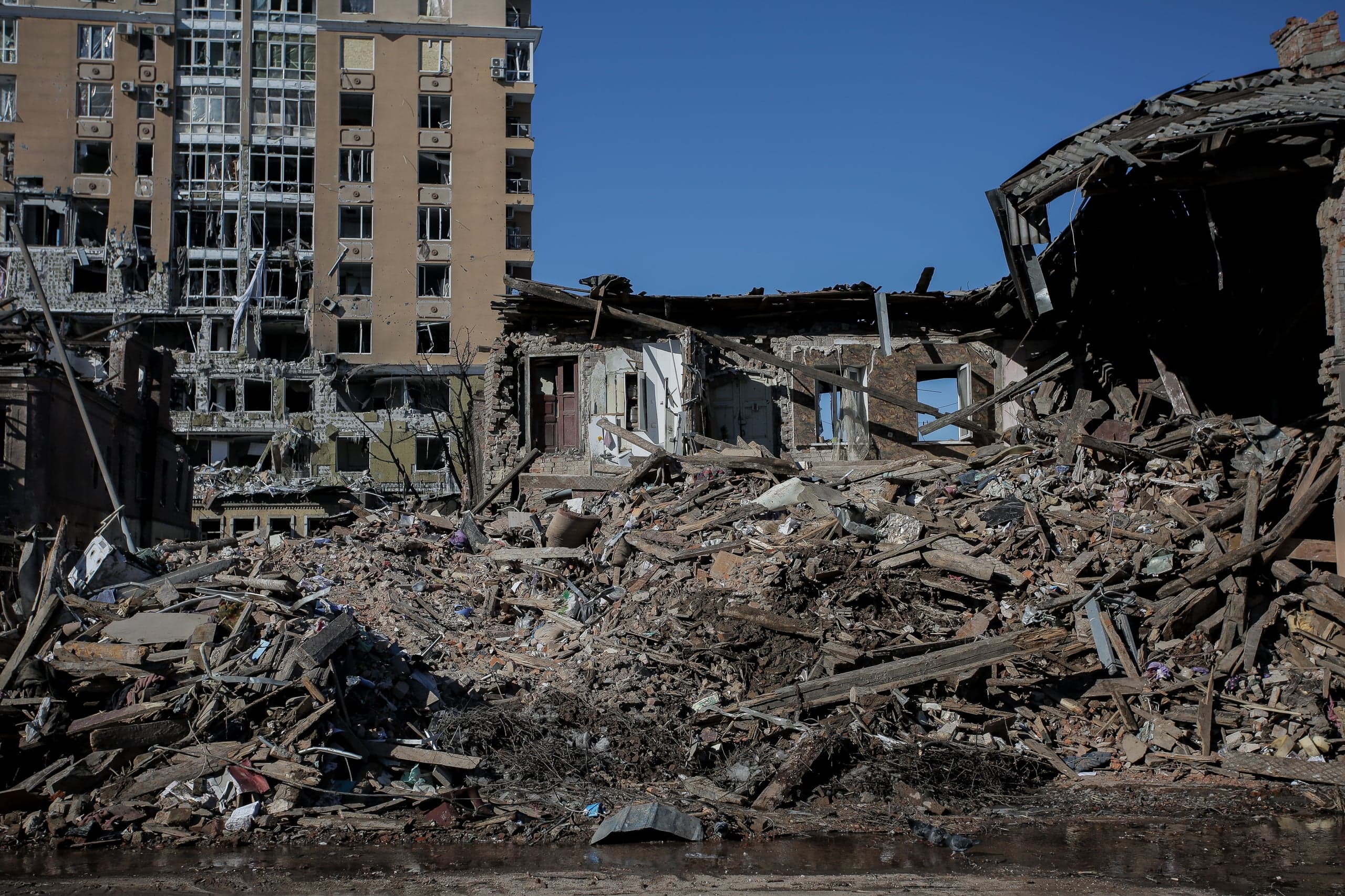 Груды строительного мусора и обломков перед руинами здания, за которыми видна поврежденная многоэтажка.