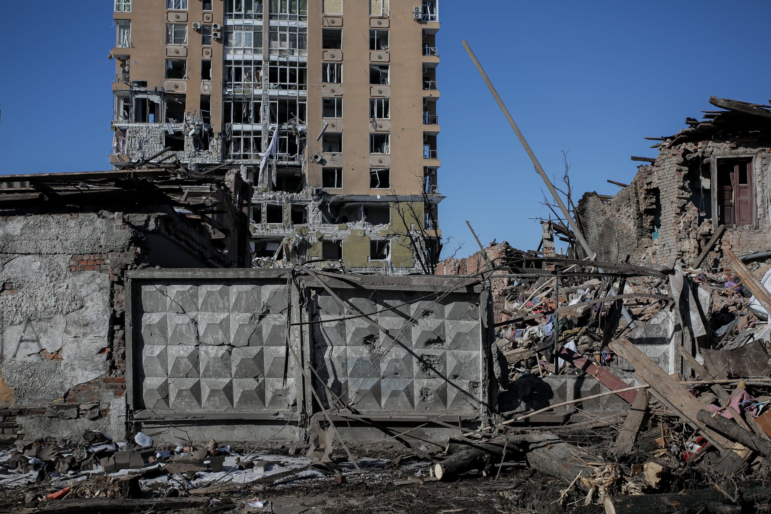 Разрушенный кирпичный забор и обломки на переднем плане, на заднем плане — многоэтажный жилой дом с выбитыми окнами и поврежденным фасадом.