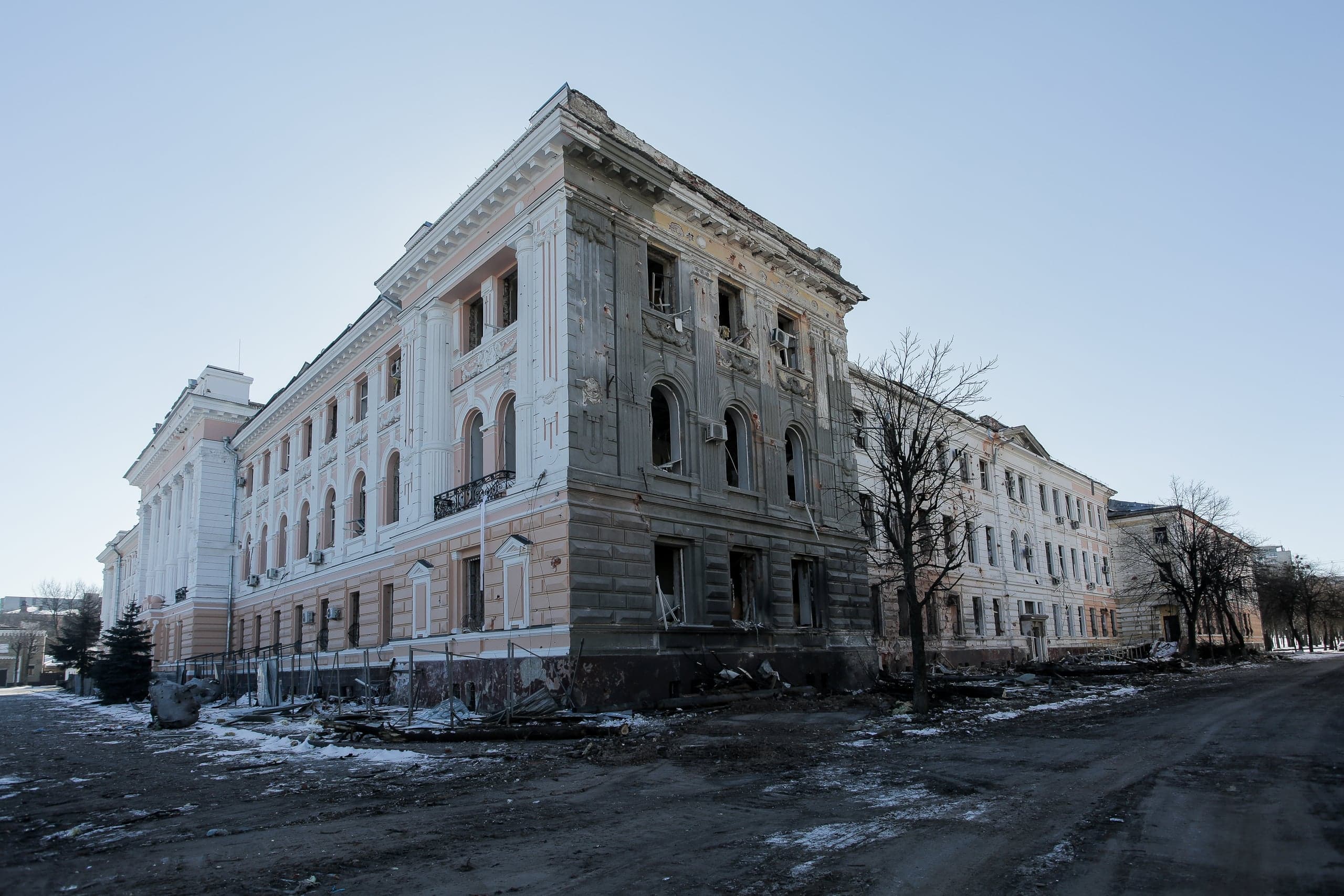 Сильно поврежденное обстрелами трехэтажное здание с классическим фасадом, выбитыми окнами и обломками на грязной дороге.