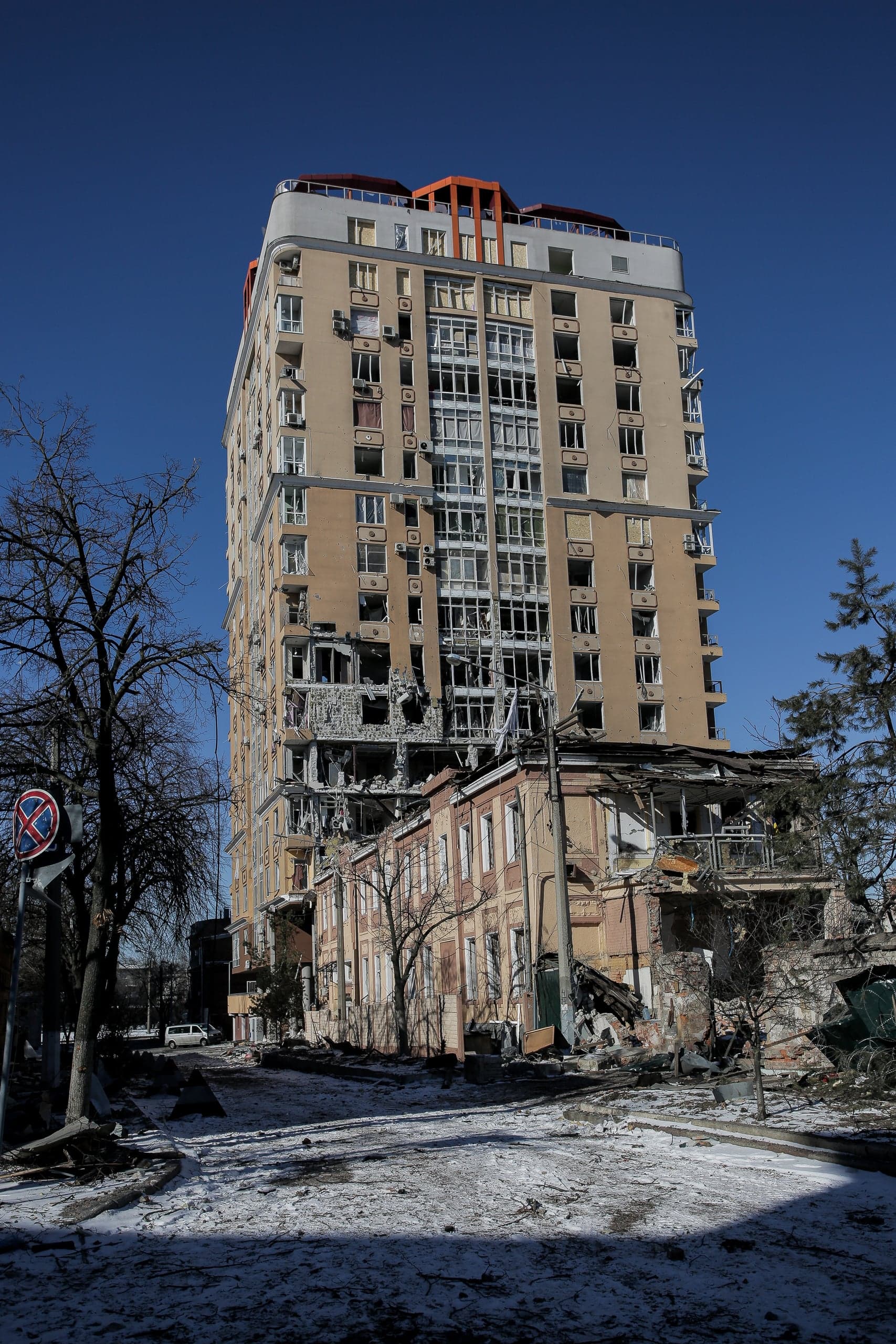 Разрушенное многоэтажное здание с выбитыми окнами и поврежденным фасадом на фоне ясного неба, рядом полуразрушенный малоэтажный дом. Снег на улице.
