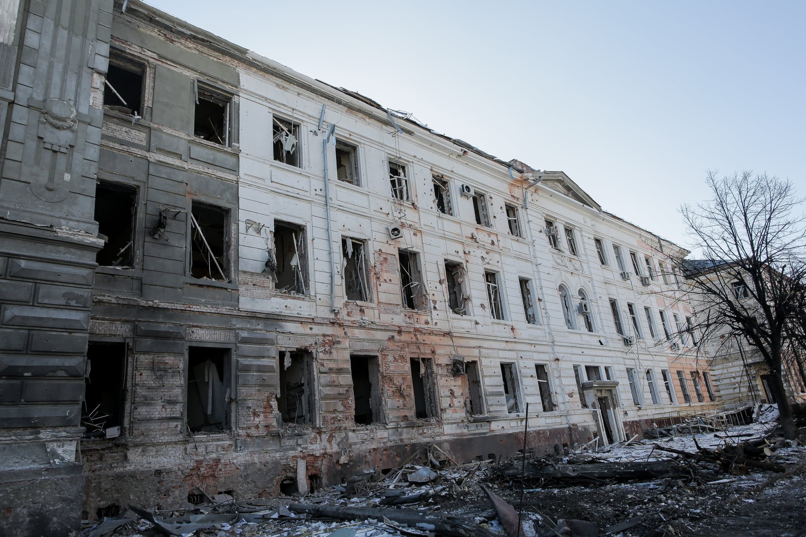 Разрушенное трехэтажное здание с выбитыми окнами, следами обстрелов на белом и сером фасаде и обломками на земле.
