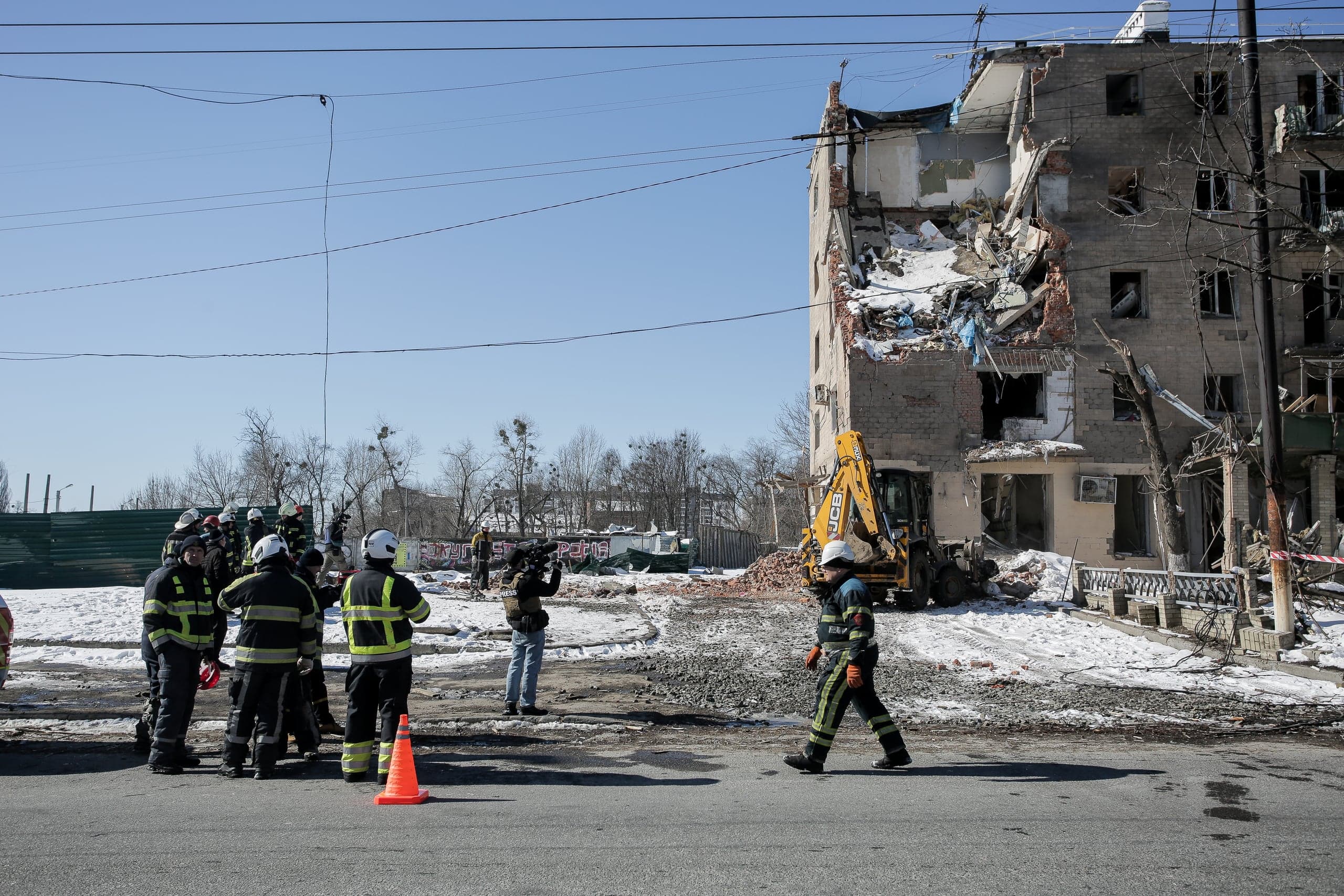 Разрушенное многоэтажное здание. Спасатели и экскаватор работают на месте обрушения на фоне голубого неба.