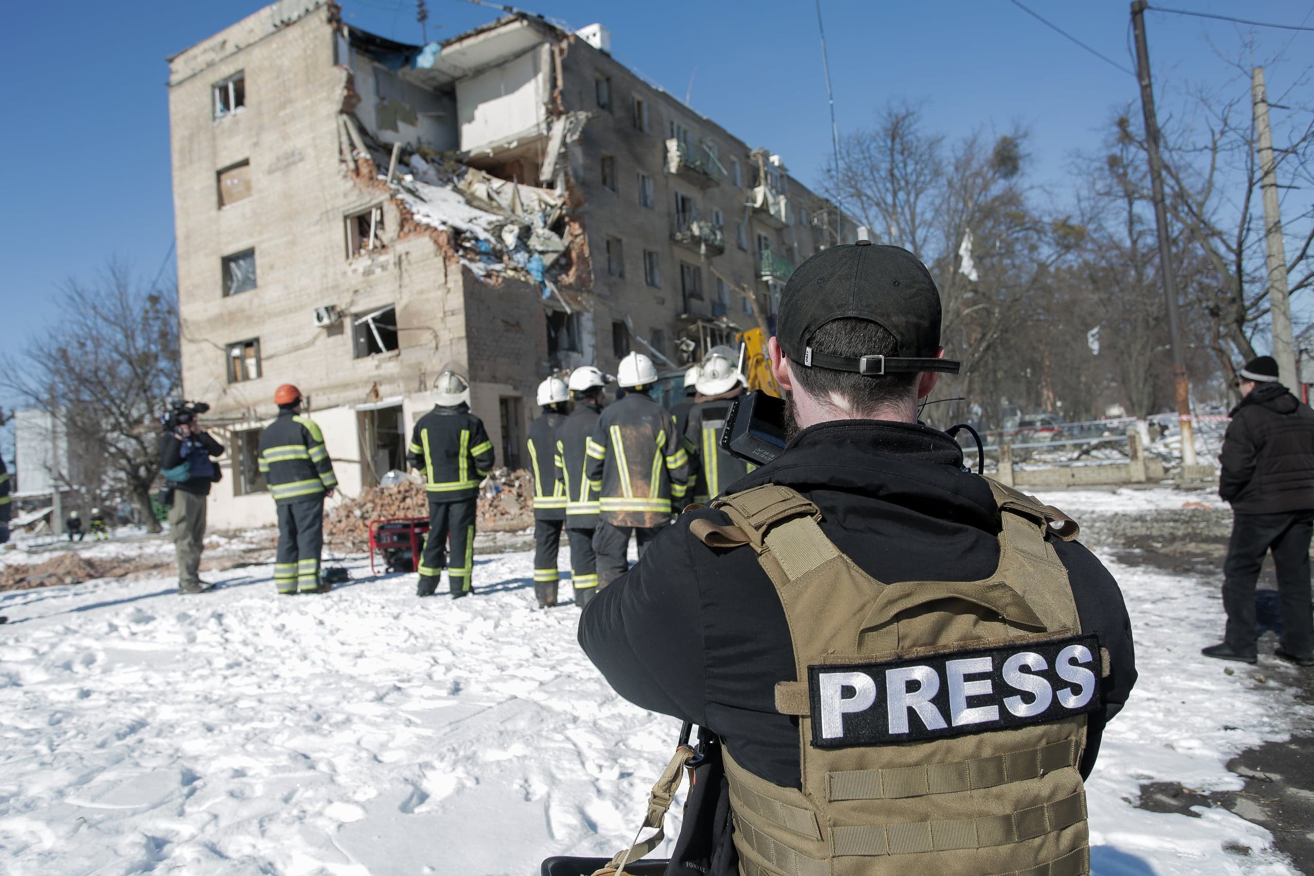 Журналист в жилете с надписью "PRESS" снимает разрушенное жилое здание и работающих перед ним спасателей на фоне снега.