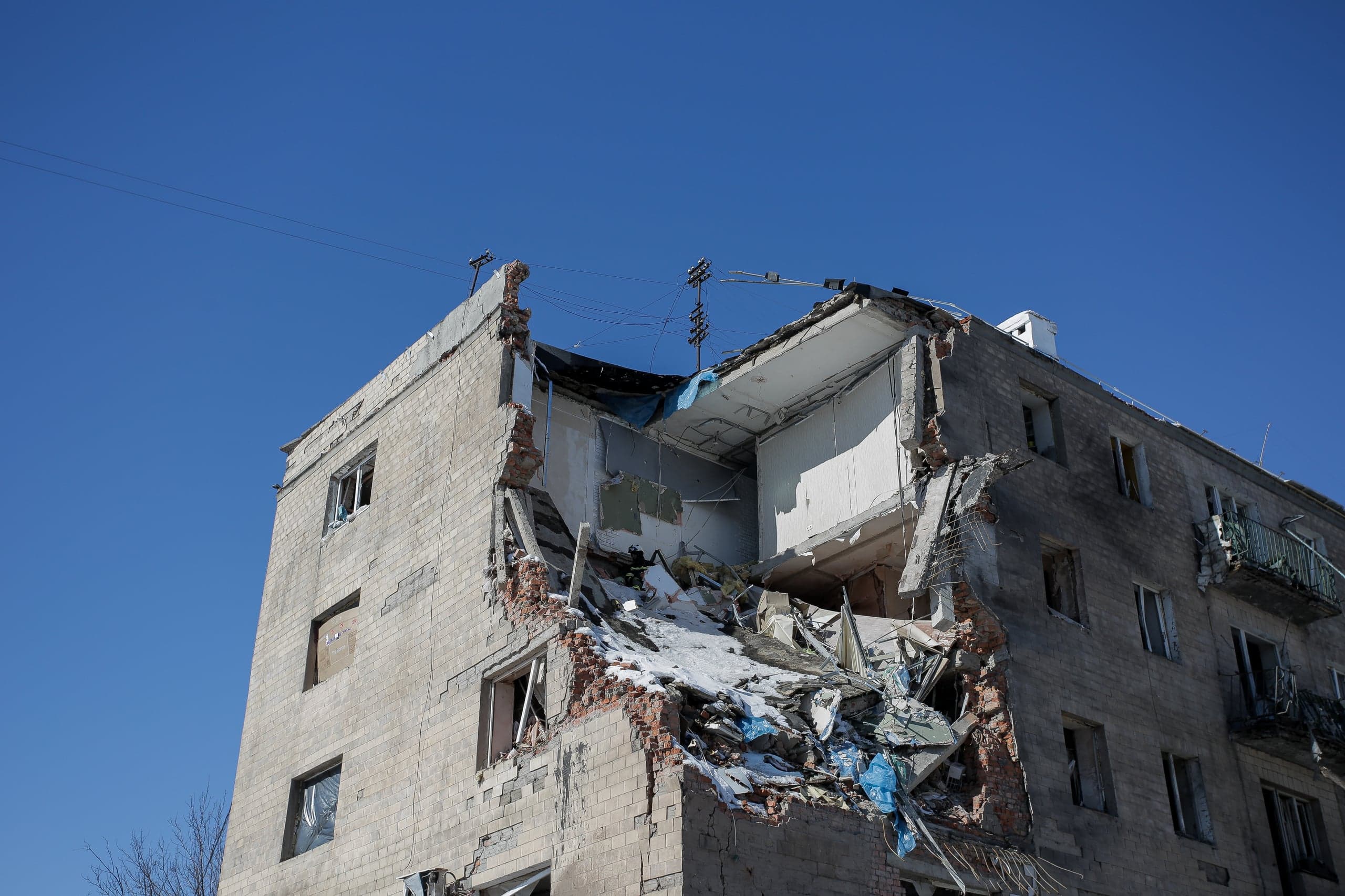 Обрушенный угол многоквартирного дома с грудой обломков и частично сохранившимся фасадом под ясным небом.