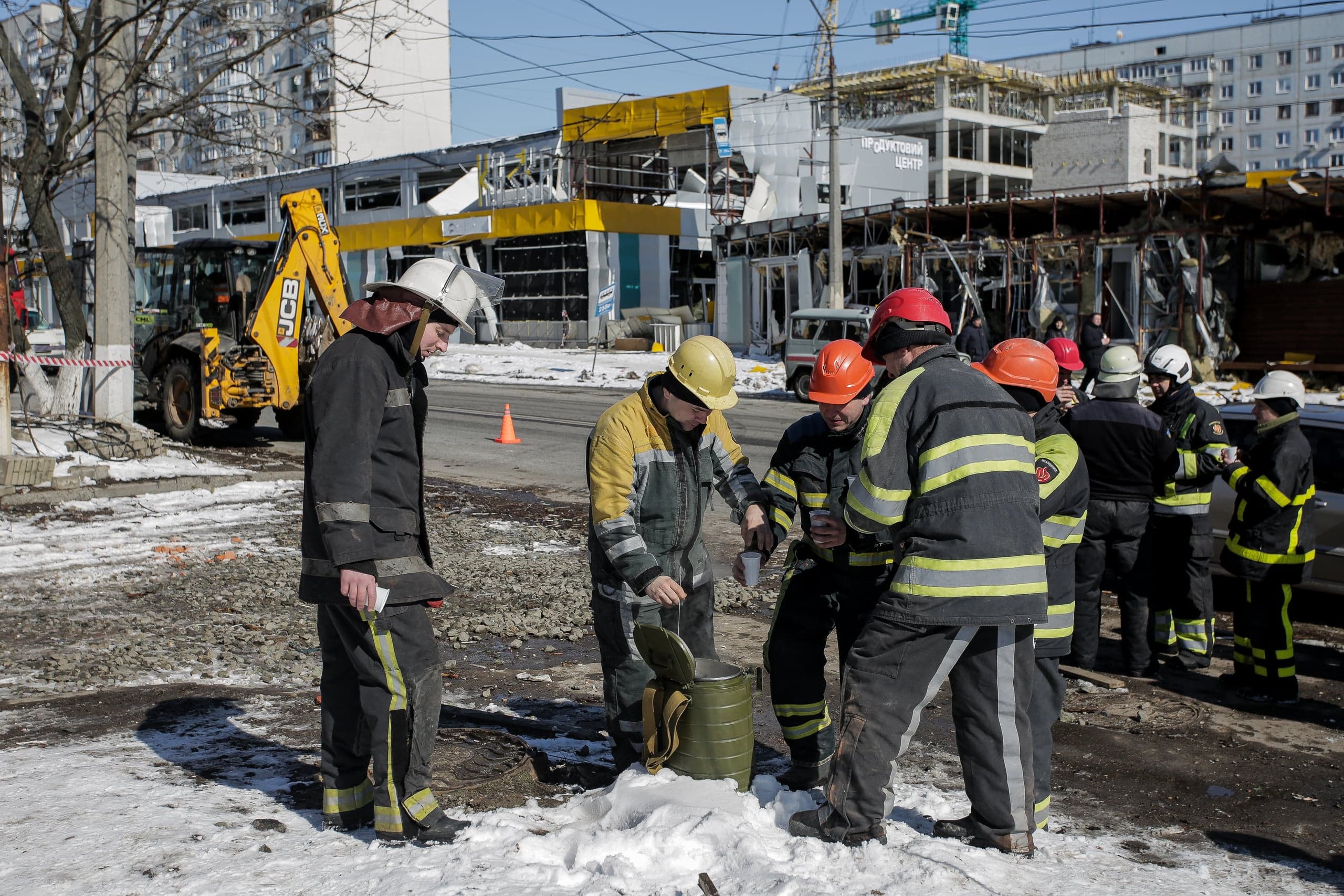 Группа спасателей и пожарных наливает горячий напиток на фоне разрушенного здания "ПРОДУКТОВИЙ ЦЕНТР" и строительной техники.