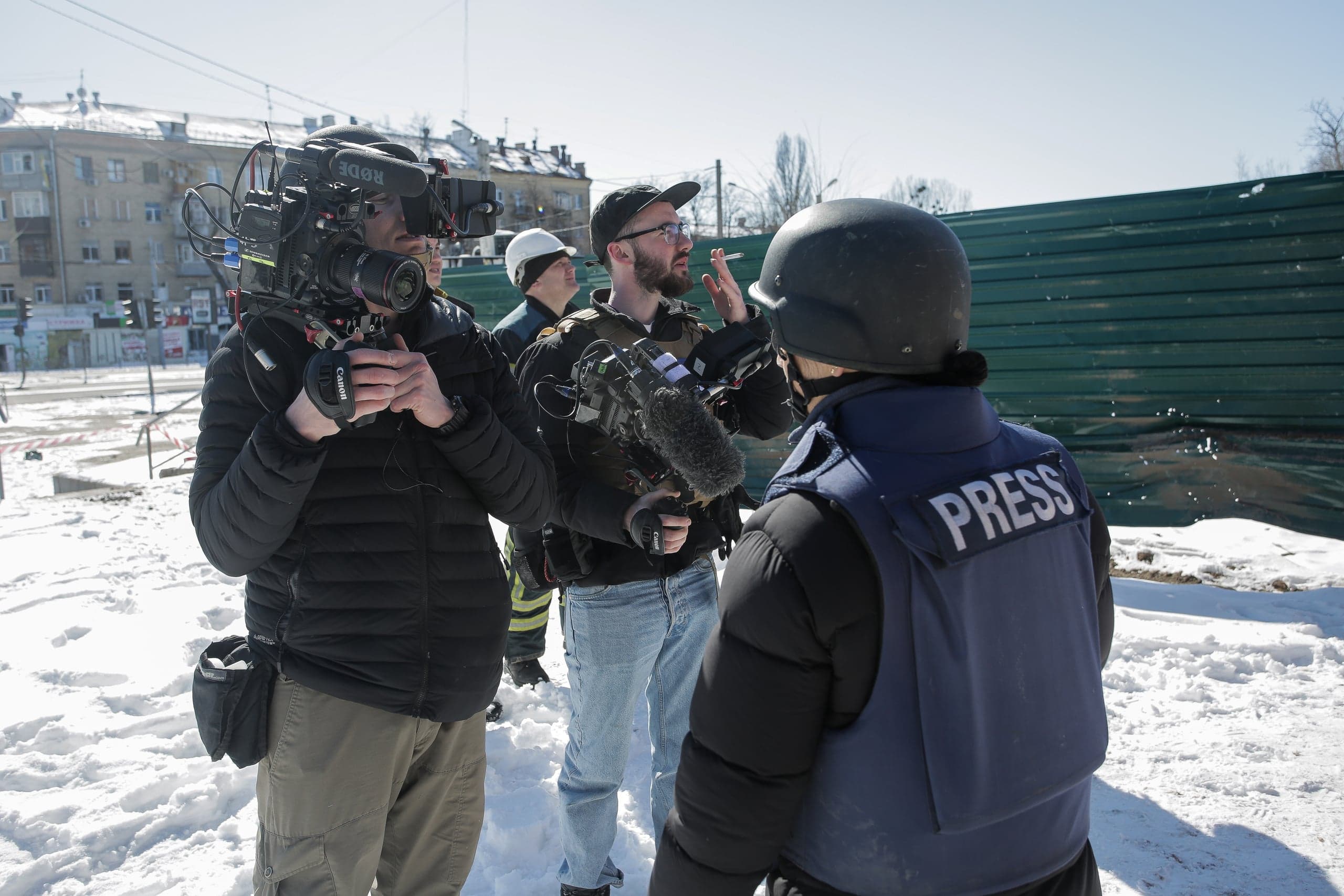 Журналисты и оператор с профессиональной камерой в зимней локации. На спине бронежилета видна надпись "PRESS".