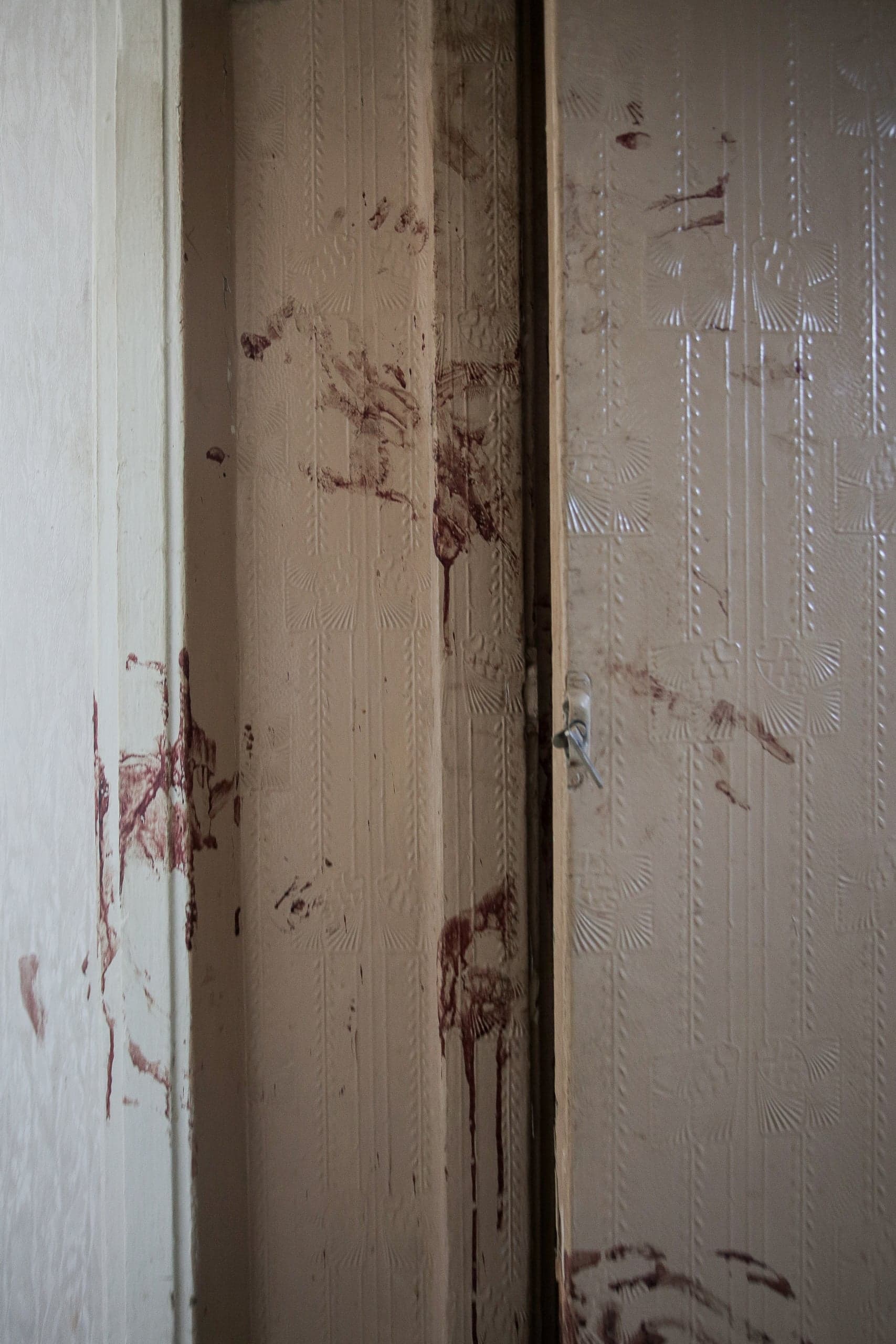 Следы крови и кровавые отпечатки рук на бежевых обоях и дверном косяке.