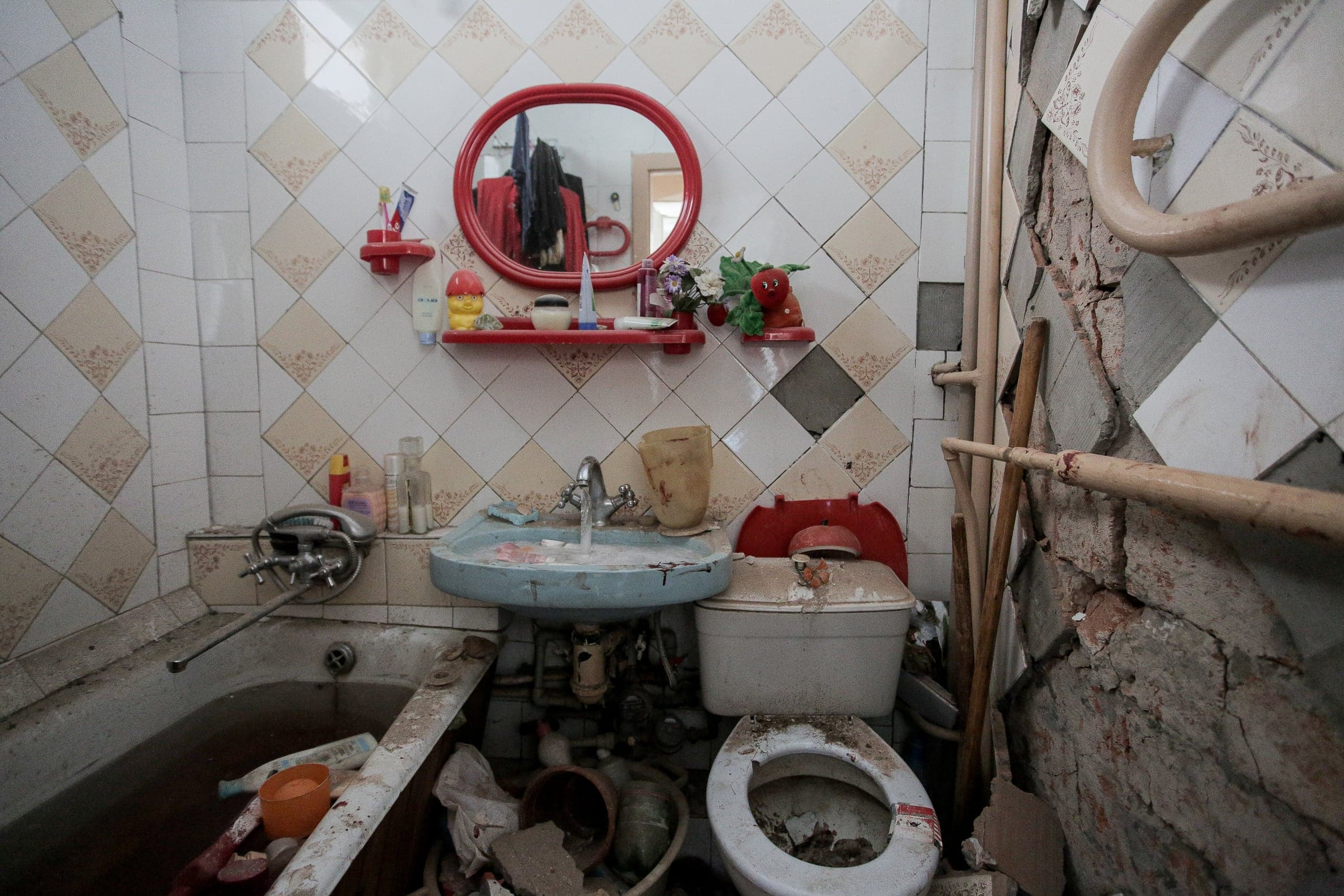 Сильно поврежденная ванная комната с грязными сантехническими приборами (ванна, раковина, унитаз). Правая стена обрушена, обнажая кирпичную кладку и трубы.