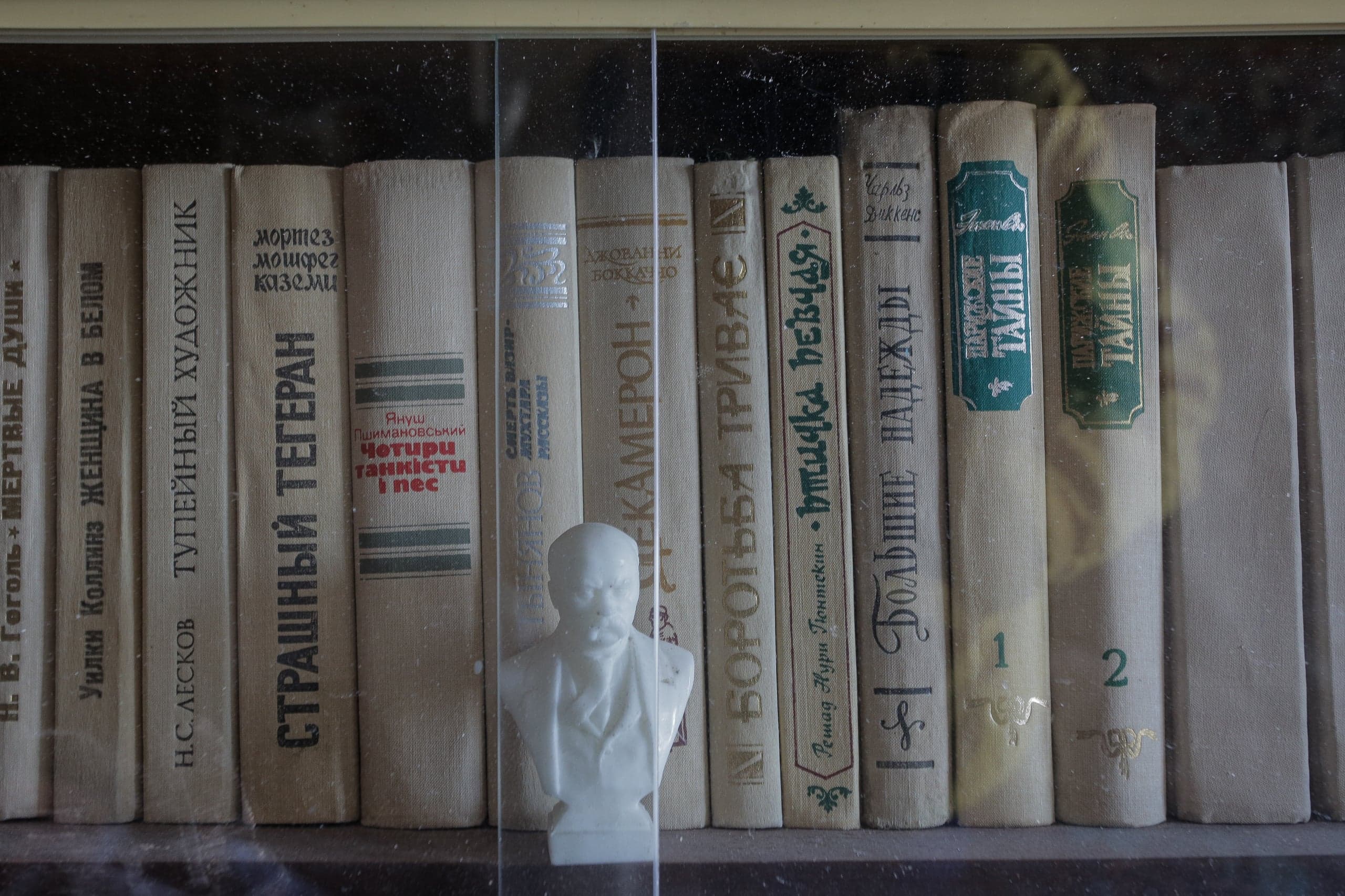 Книжная полка с рядами светлых книг в одинаковых переплетах и небольшим белым бюстом Ленина. Среди названий: "СТРАШНЫЙ ТЕГЕРАН", "ТУПЕЙНЫЙ ХУДОЖНИК", "БОЛЬШИЕ НАДЕЖДЫ", "МОРСКИЕ ТАЙНЫ".