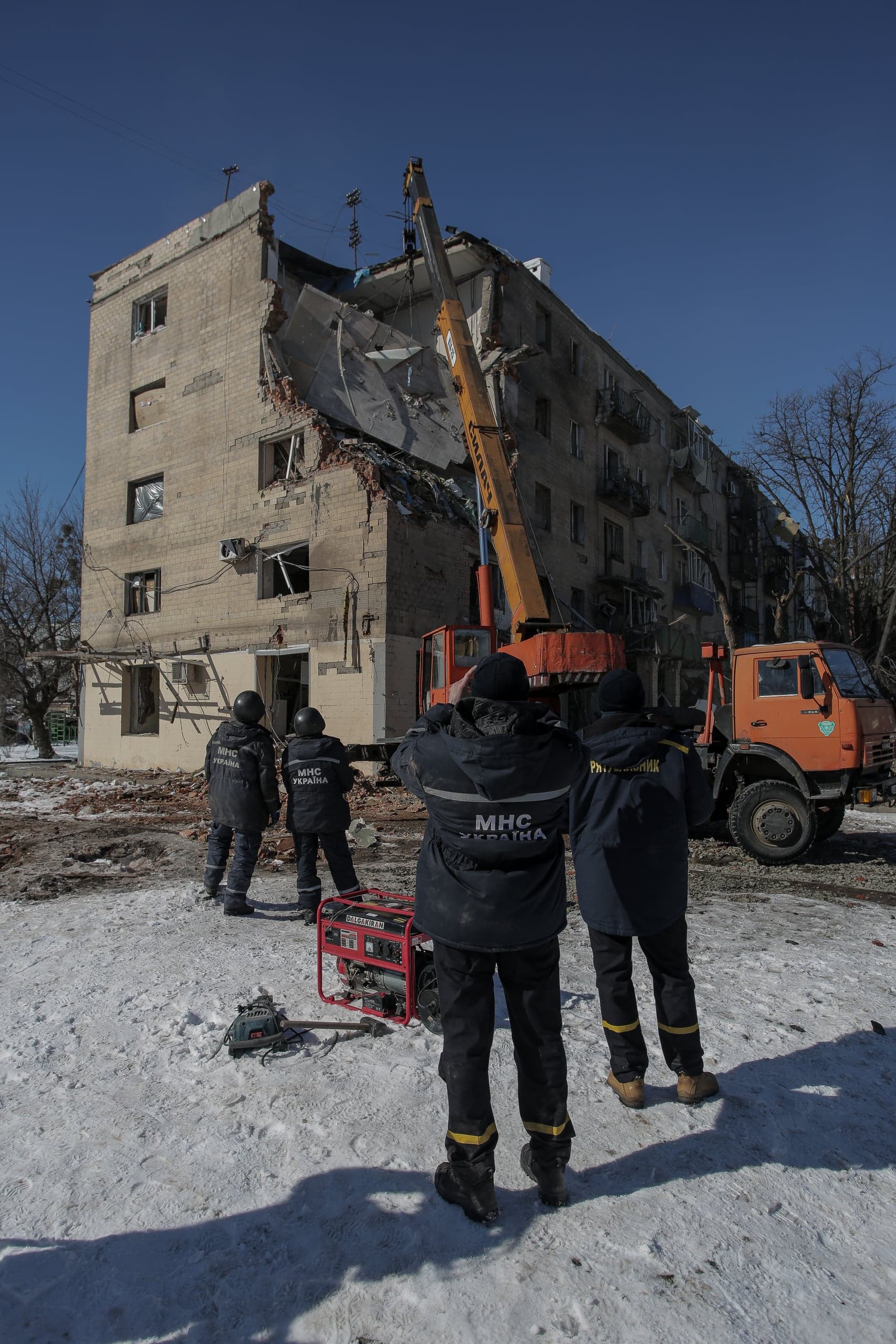 Спасатели "МНС УКРАЇНА" и "РЯТІВНИК" работают у частично разрушенного многоквартирного дома. Кран разбирает завалы на фоне снега и синего неба.