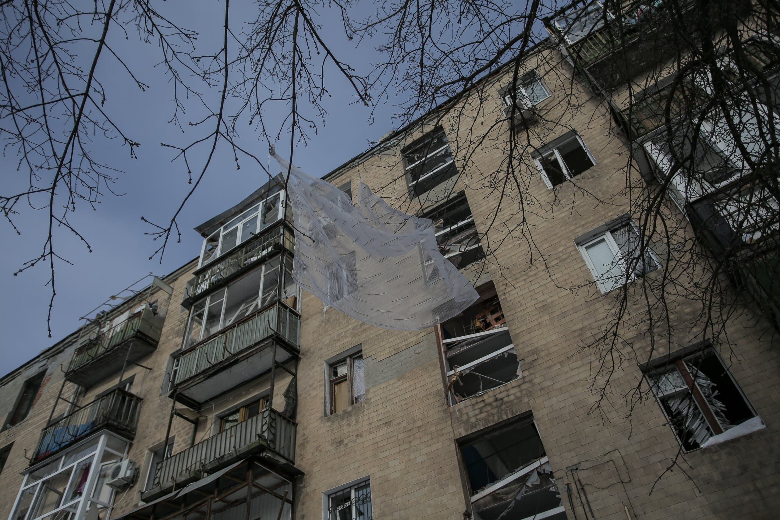 Фасад многоквартирного дома с разбитыми окнами. Кусок прозрачного полиэтилена висит на голых ветках на фоне пасмурного неба.