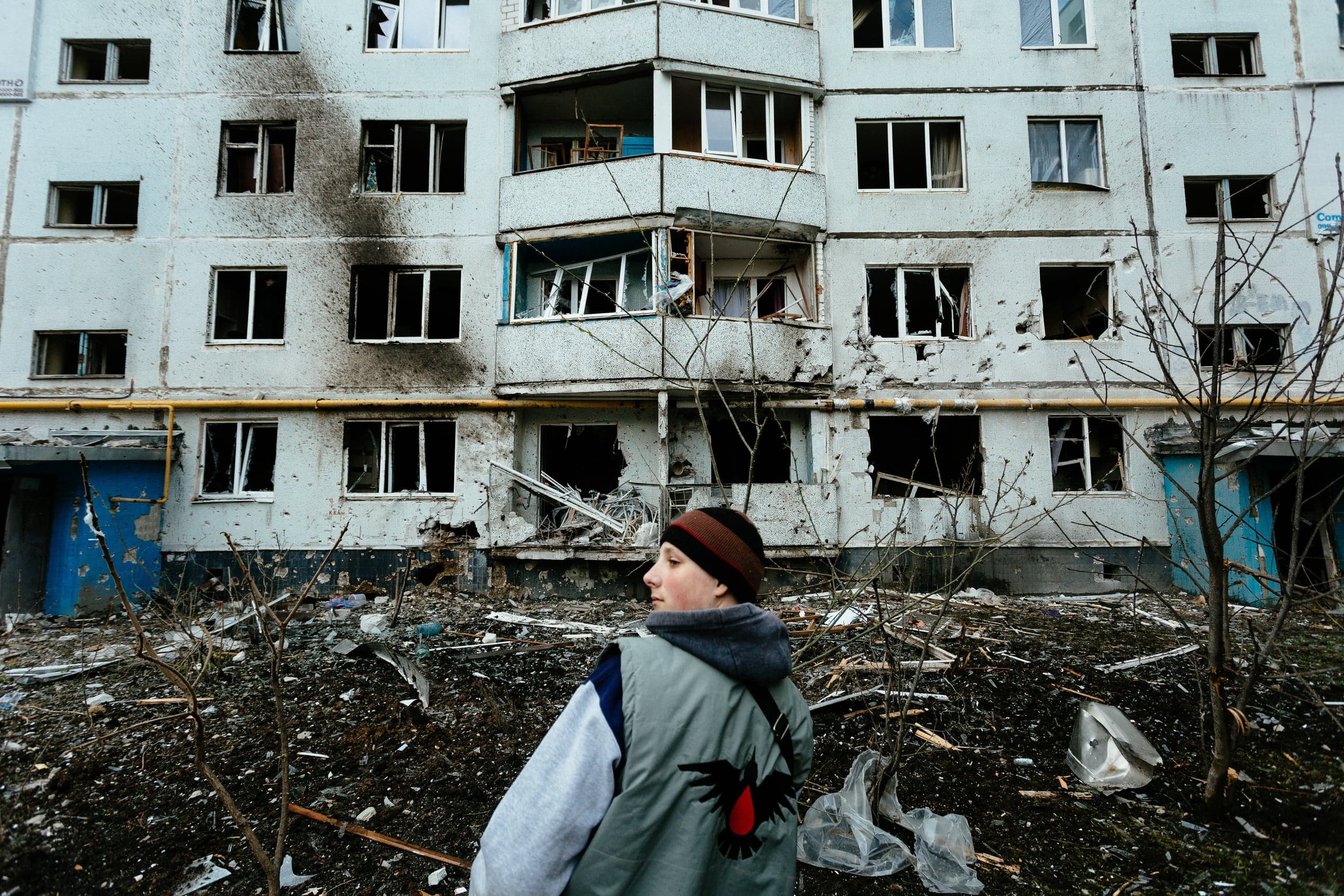 Разрушенный многоквартирный дом с выбитыми окнами и следами гари. На переднем плане человек со спины, стоящий среди обломков.
