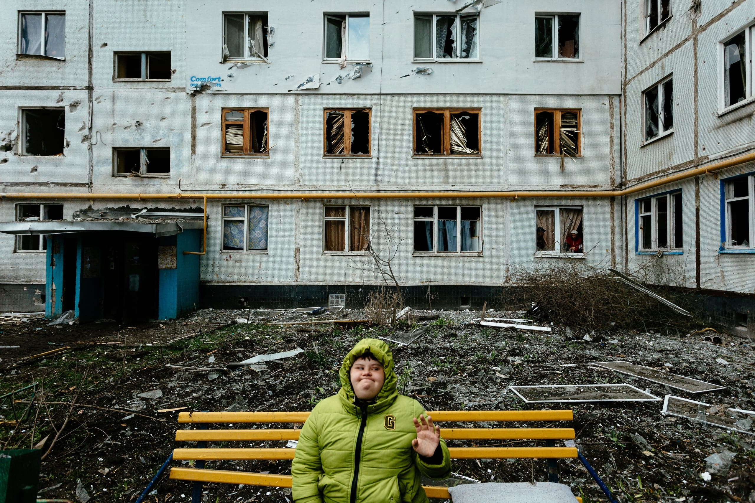 Молодой человек в салатовой куртке сидит на скамейке перед поврежденным обстрелами жилым домом с выбитыми окнами, двор завален обломками.