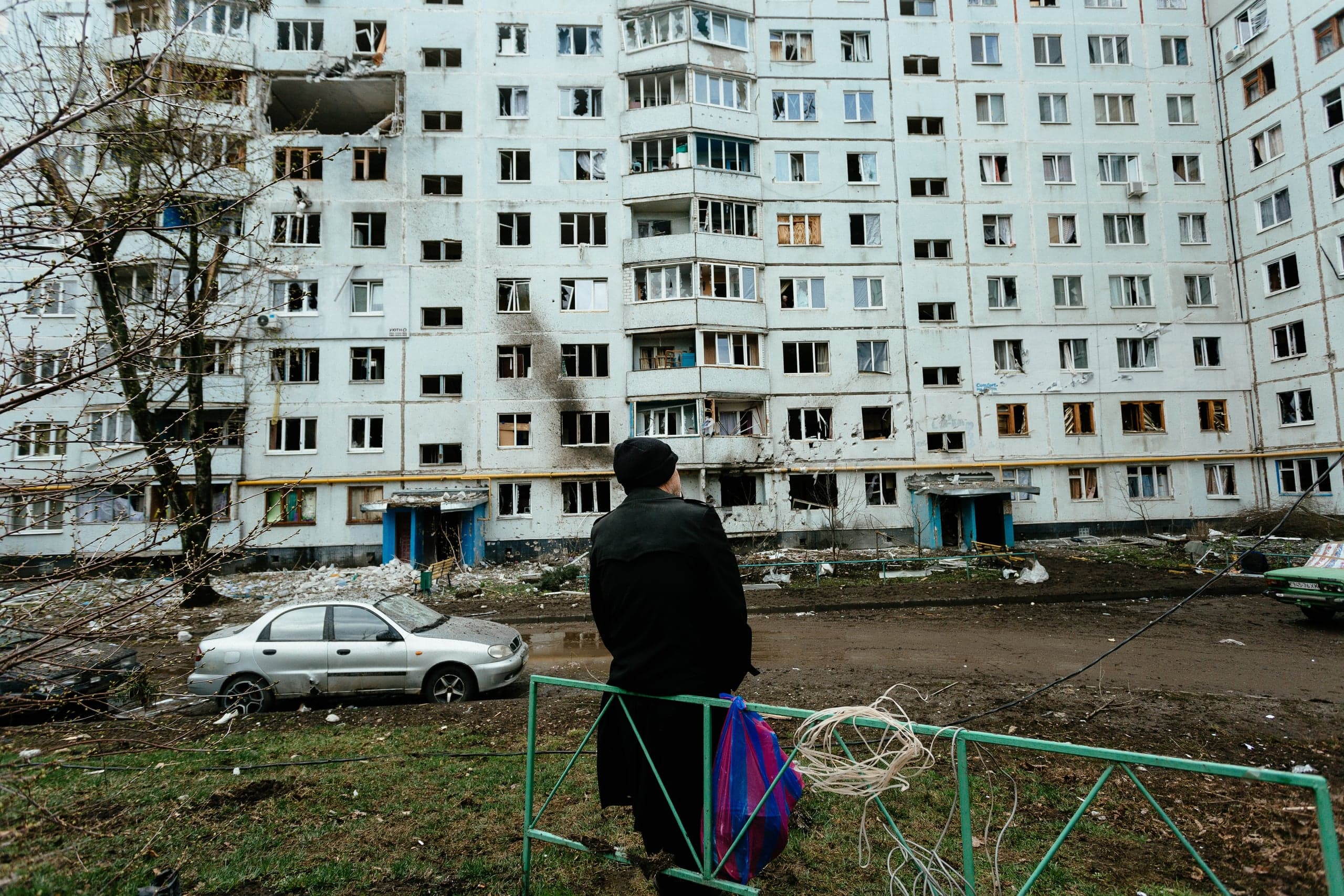 Человек стоит перед сильно поврежденным обстрелом многоэтажным жилым домом с разбитыми окнами и проломом в стене.